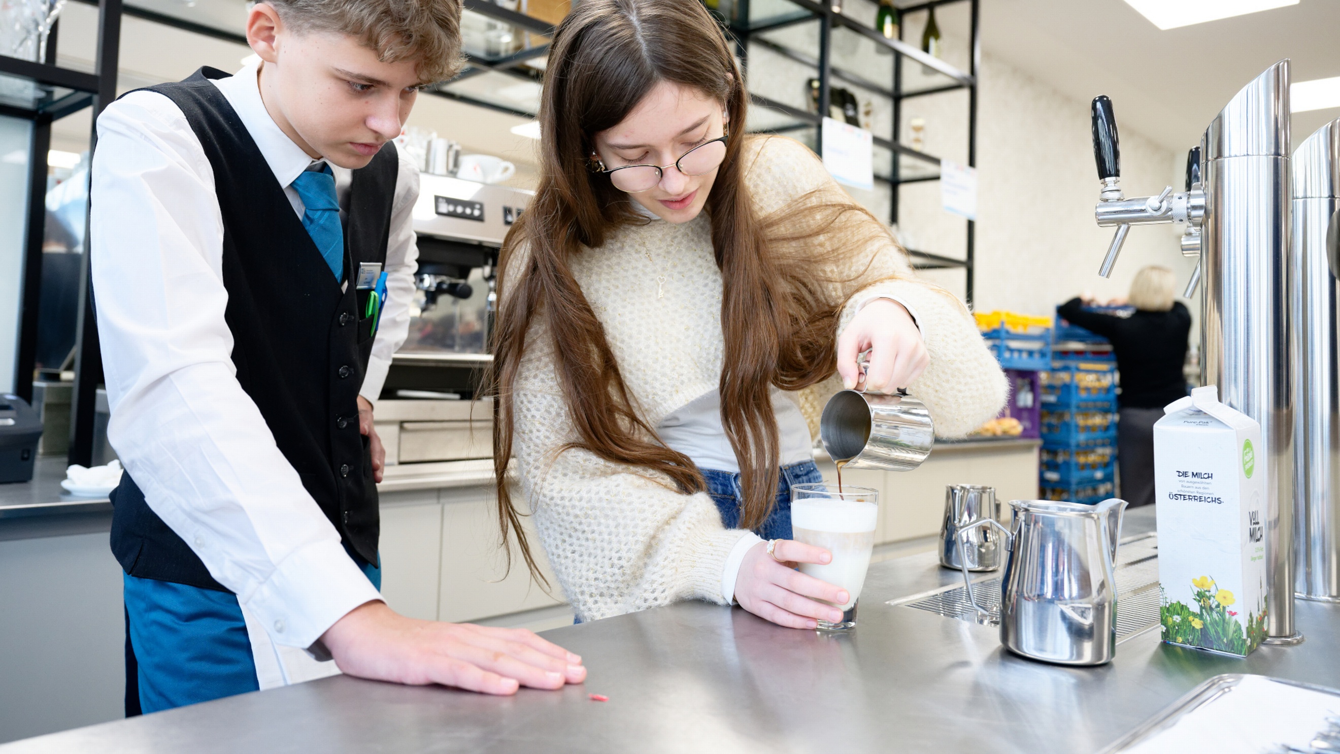 Zwei Jugendliche stehen in einer Gastroküche. Die rechte Person leert aus einer Milchkanne Kaffee in ein Glas mit Milchschaum. Die Person daneben schaut zu und trägt eine dunkle Weste sowie eine Krawatte. Auf dem Tresen ist ein Zapfhahn