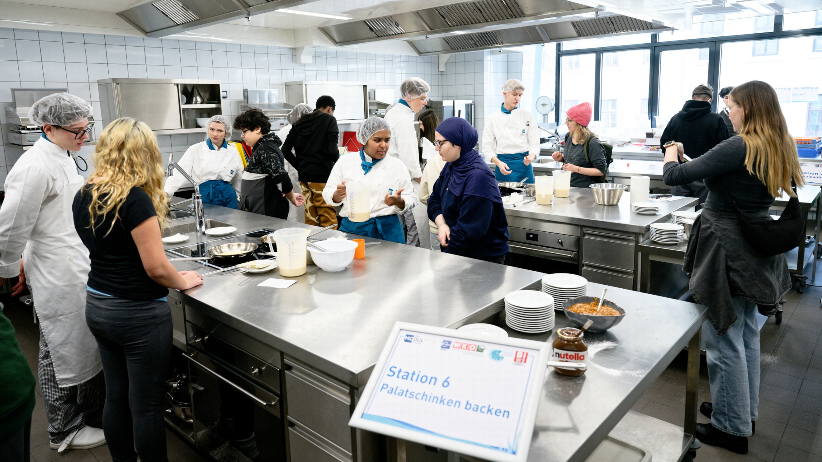  In einer Gastronomieküche stehen an verschiedenen Stationen jeweils eine Person mit Haarnetz und Schürze sowie eine jugendliche Person. Rechts vorne steht auf einem Zettel Station 6, Palatschinken backen. Daneben stehen mehrere Stapel Teller