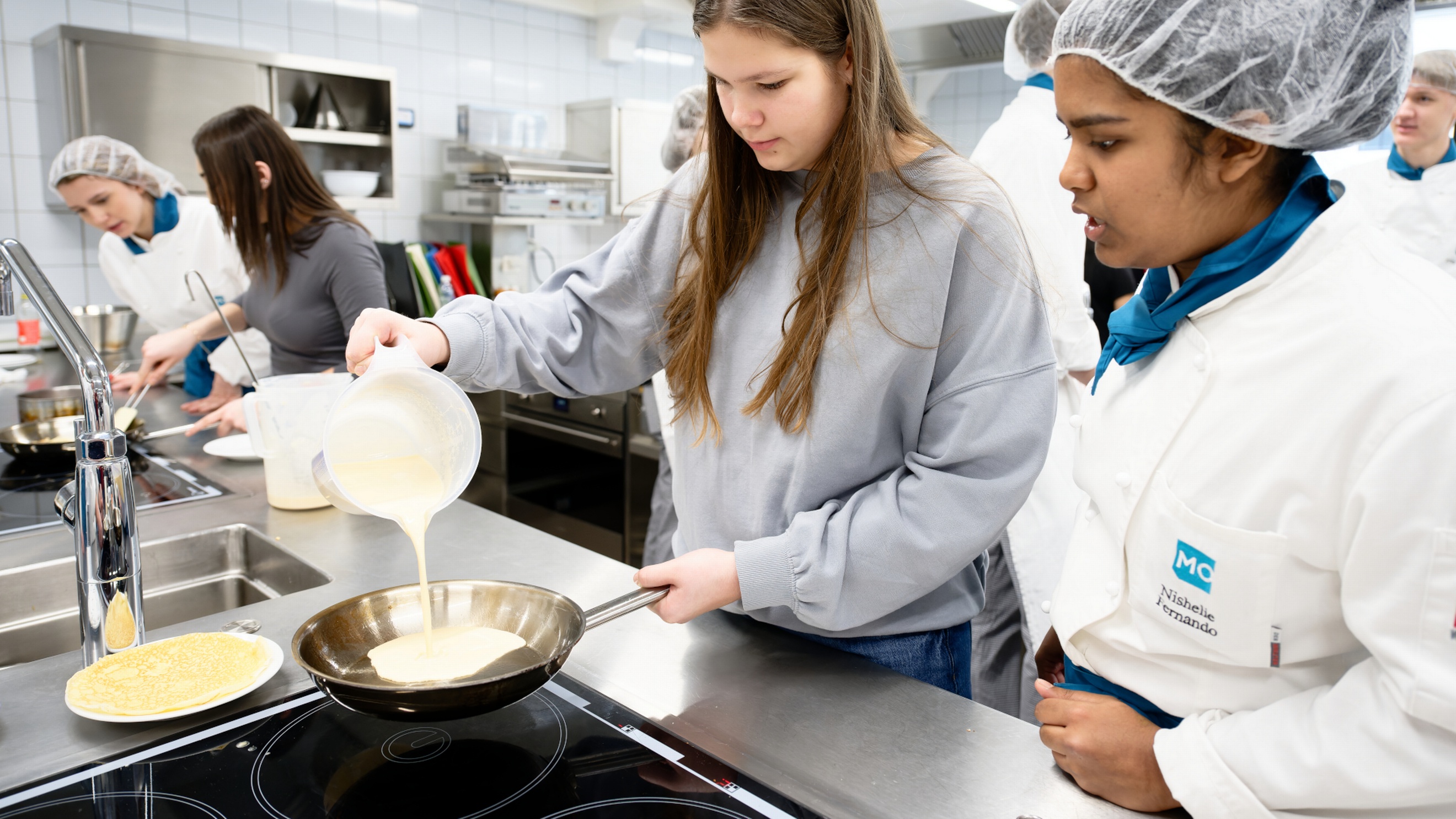 Eine Person mit Haarnetz und Schürze sowie eine Jugendliche stehen in einer Gastronomieküche neben einer Herdplatte. Die jugendliche Person hält eine Pfanne, in die sie aus einer Schüssel eine helle Flüssigkeit kippt