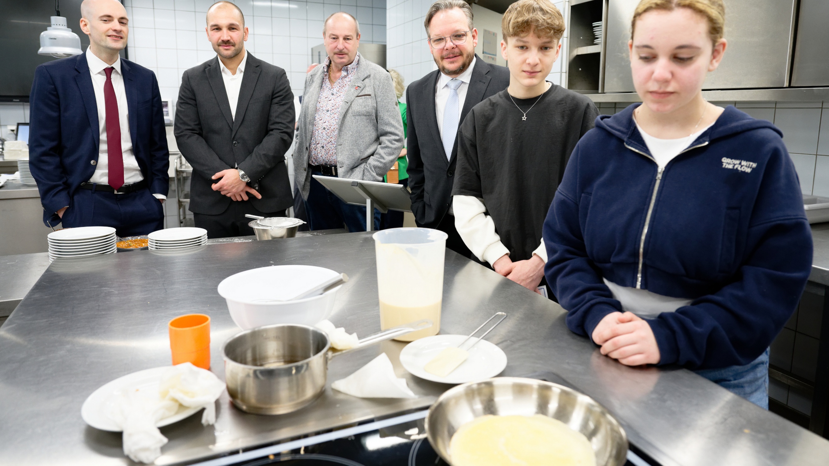 Neben einem Arbeitstisch in einer Gastronomieküche stehen vier erwachsene Personen sowie zwei Jugendliche. Auf dem Arbeitstisch sind Schüsseln, Teller, ein Topf und eine Pfanne mit einem hellen Teig
