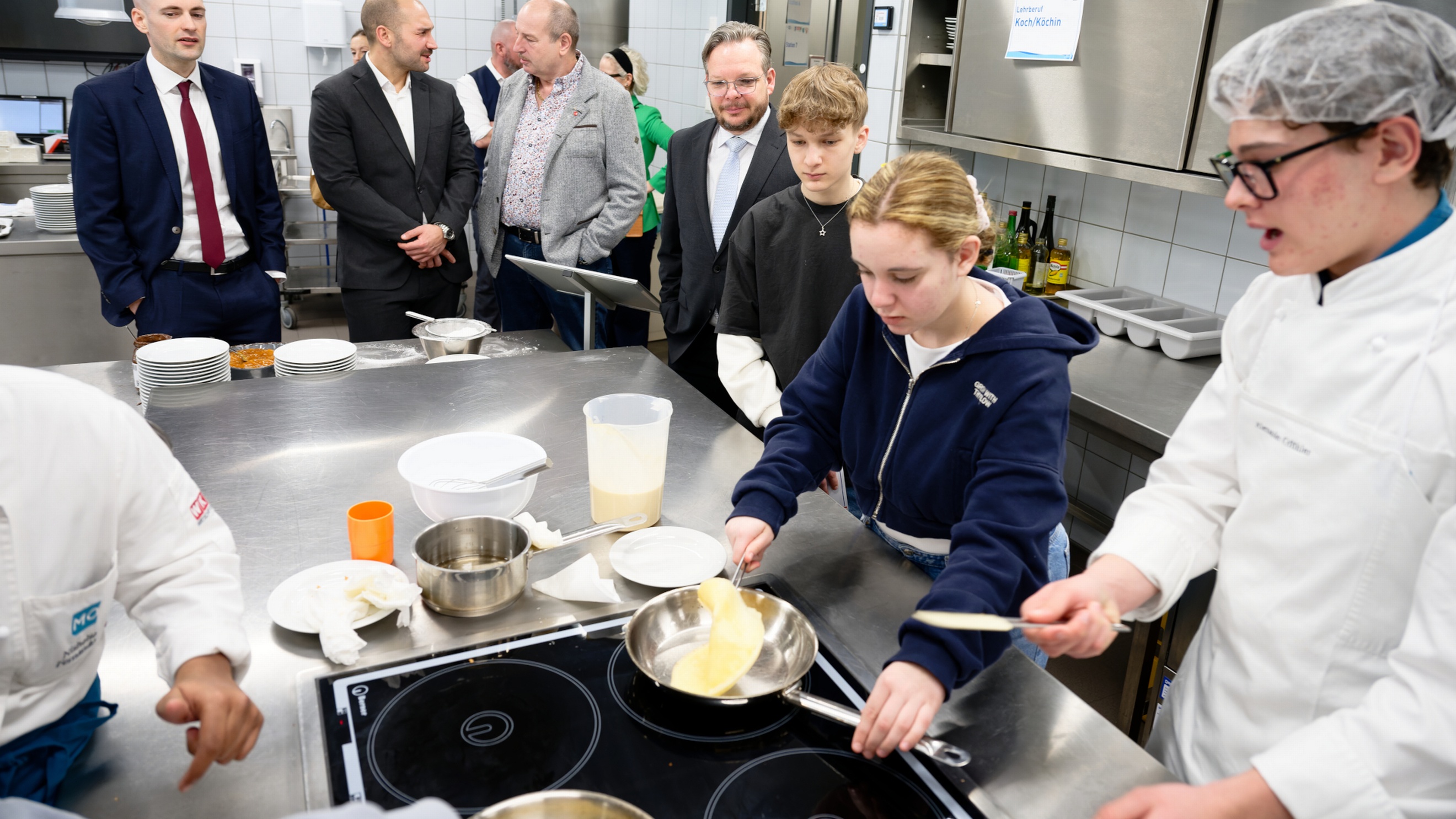 Neben einem Arbeitstisch in einer Gastronomieküche stehen vier erwachsene Personen. Neben ihnen sind zwei Jugendliche sowie eine Person mit Haube und Schürze. Auf dem Arbeitstisch ist eine Herdplatte, auf der eine Pfanne steht. Eine der jugendlichen Personen kocht darin