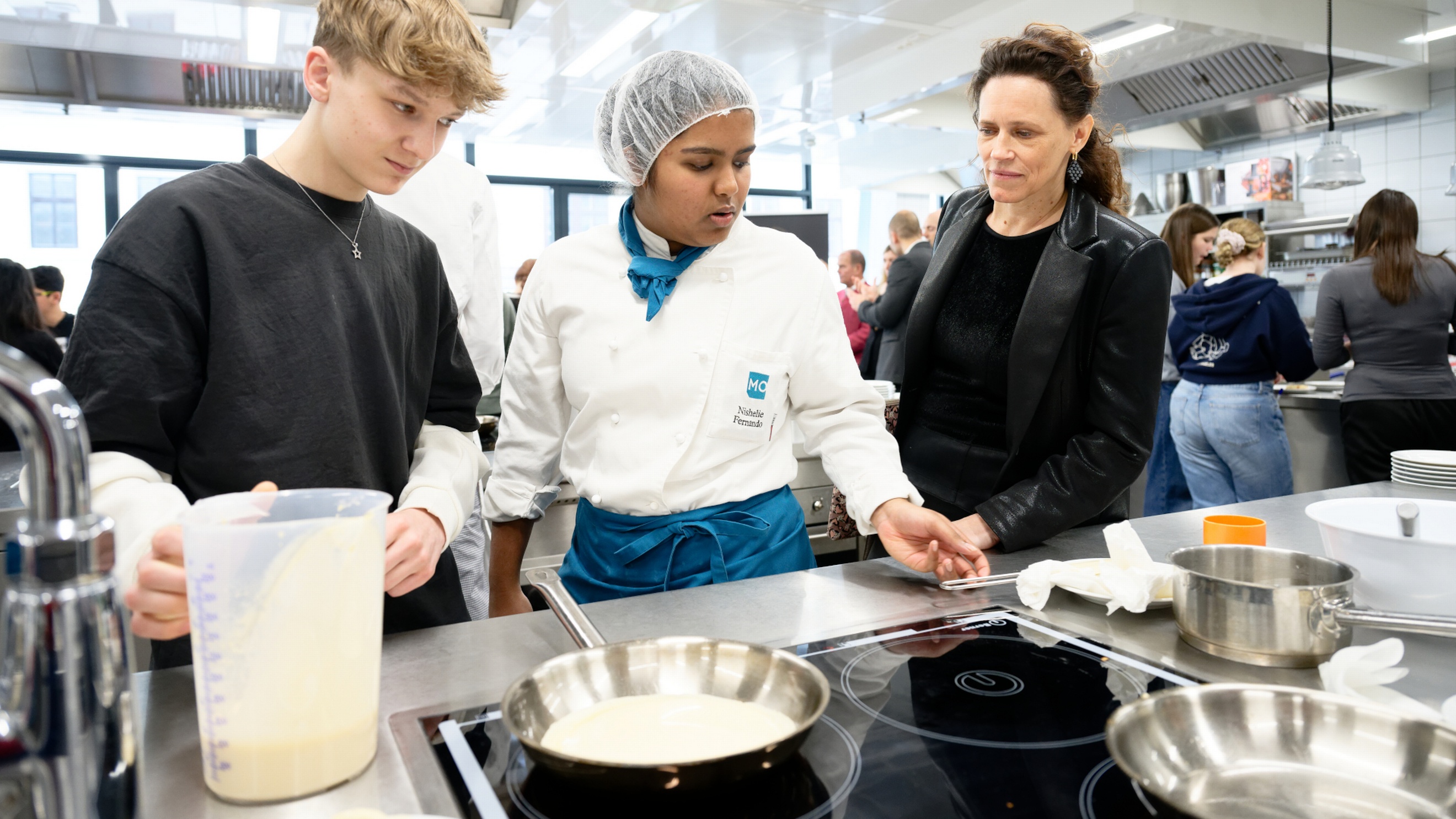 In einer Gastronomieküche stehen neben einer Herdplatte eine jugendliche Person, eine Person mit Haarnetz und Schürze sowie eine erwachsene Person. Auf der Herdplatte steht eine Pfanne. Die jugendliche Person hält ein Plastikgefäß, in dem eine helle Flüssigkeit ist