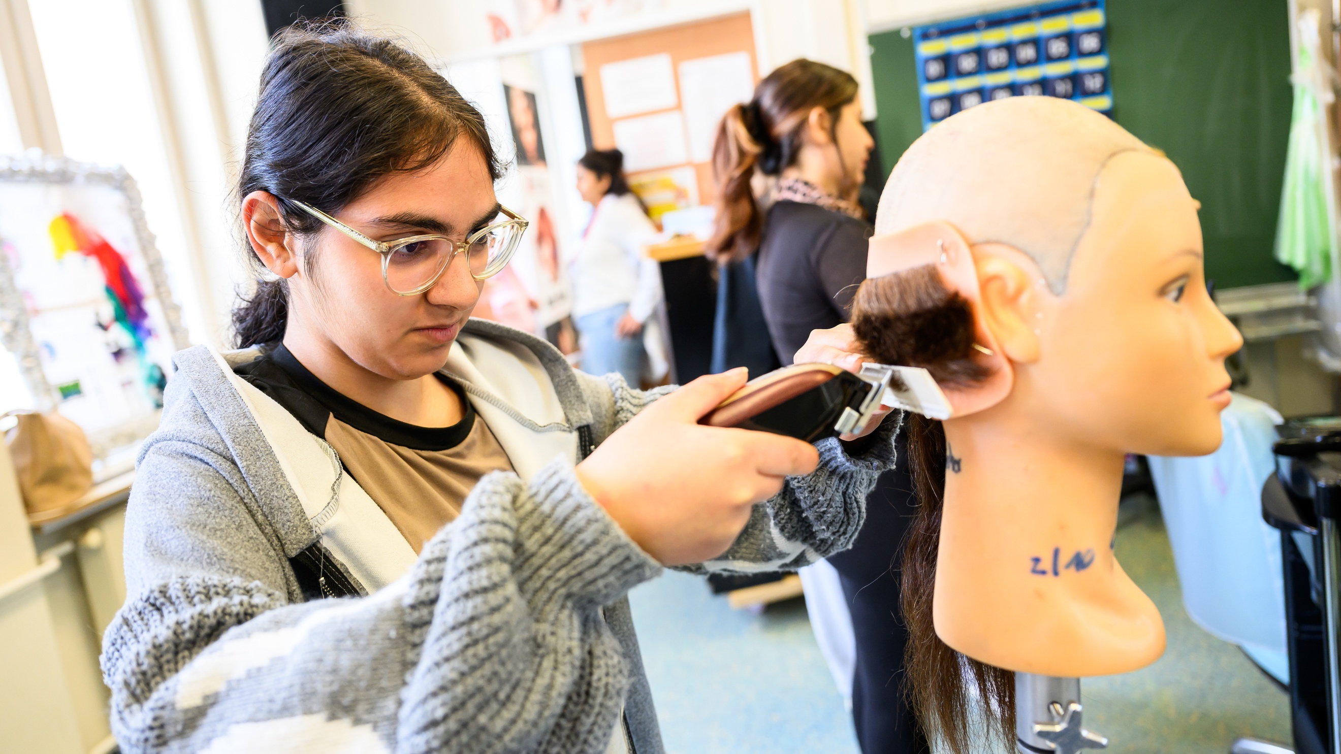 Seitliche Aufnahme des Hinterkopfes einer Modepuppe, die oben keine Haare hat. Im unteren Bereich sind Haare. Eine Person hält einen Rasierer und einen Kamm auf die Haare