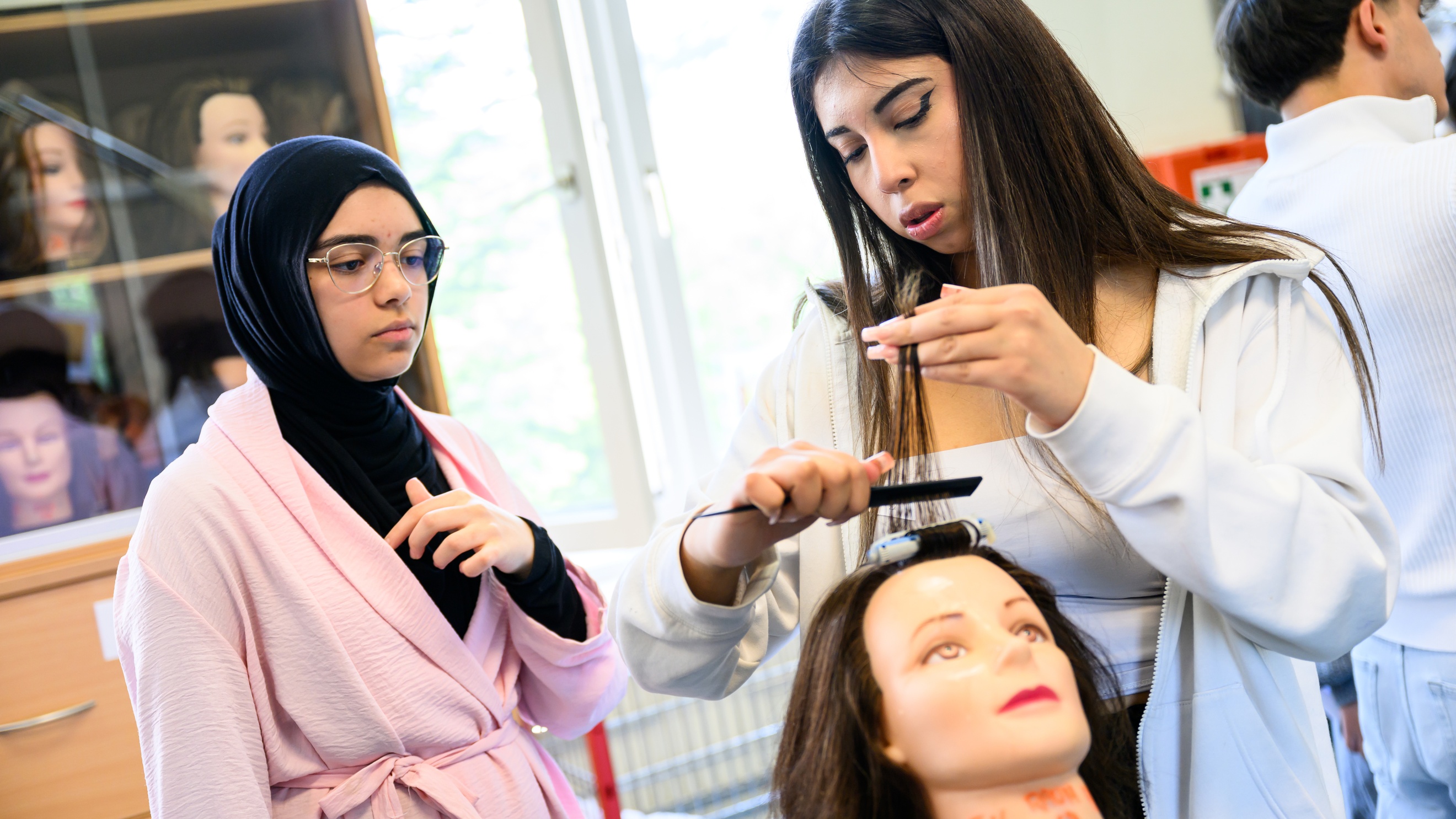 Eine Person hält die Haarsträhne eines Modepuppenkopfes mit der linken Hand. Mit der rechten Hand kämmt sie die Strähne. Neben ihr steht eine jugendliche Person, die zusieht