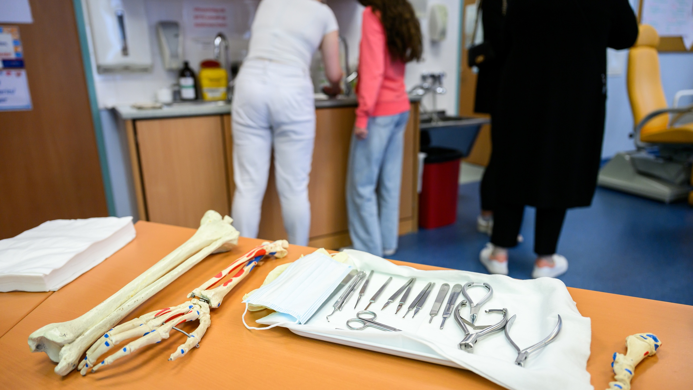Im Vordergrund liegen auf einem Tisch verschiedene Utensilien wie Scheren oder Pinzetten. Daneben liegen Modelle von Händen und Knochen. Im Hintergrund stehen Personen herum