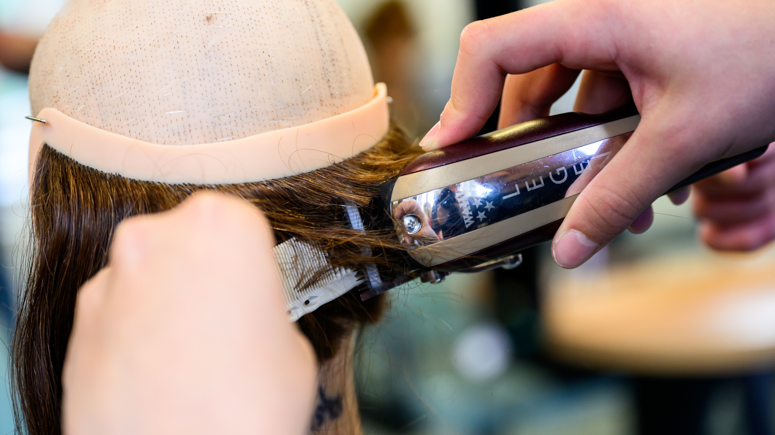 Nahaufnahme des Hinterkopfes einer Modepuppe, die oben keine Haare hat. Im unteren Bereich sind Haare. Eine Person hält einen Rasierer und einen Kamm auf die Haare