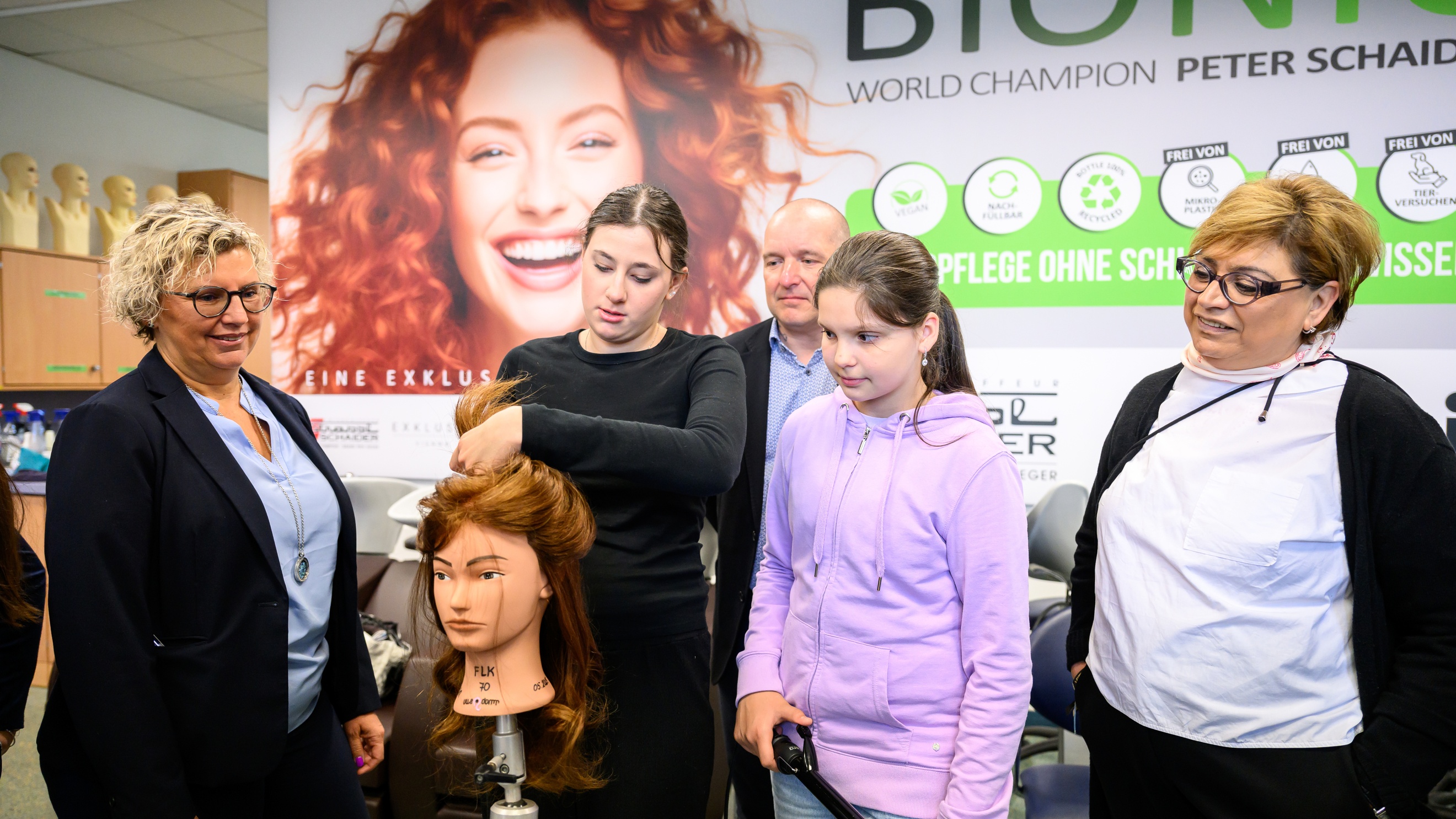Ein Modepuppenkopf mit langem Haar ist auf einer Metallstange montiert. Dahinter steht eine jugendliche Person, die der Puppe einen Zopf macht. Neben ihr stehen eine weitere jugendliche Person und drei erwachsene Personen, die zusehen