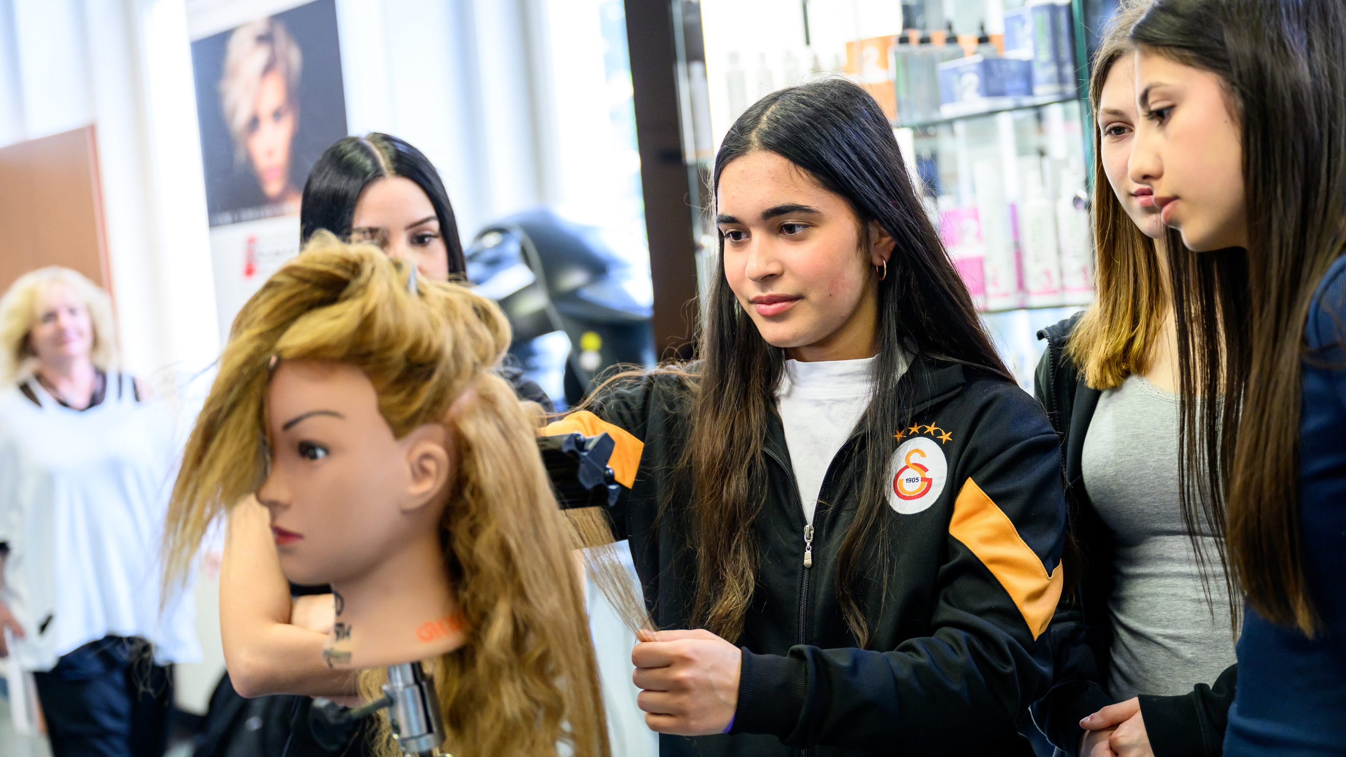 Ein Modepuppenkopf mit langem Haar ist auf einer Metallstange montiert. Dahinter steht eine jugendliche Person, die die Haare der Puppe glättet. Daneben stehen weitere jugendliche Personen