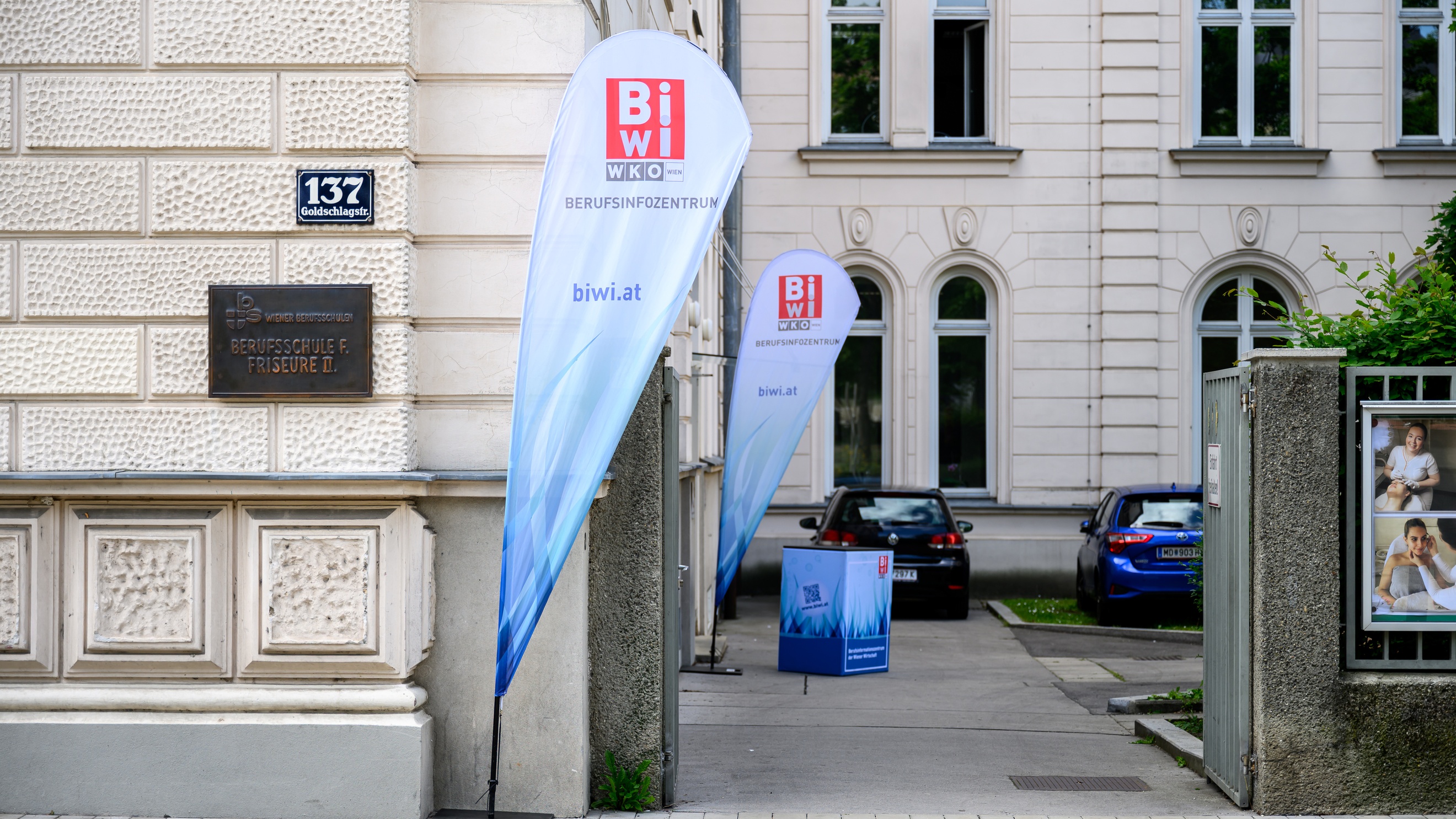 Vor der Fassade eines Hauses steht ein Werbebanner, auf dem Berufsinfozentrum und biwi.at steht. Auf der Fassade ist eine Plakette, auf der Berufsschule für Friseure steht