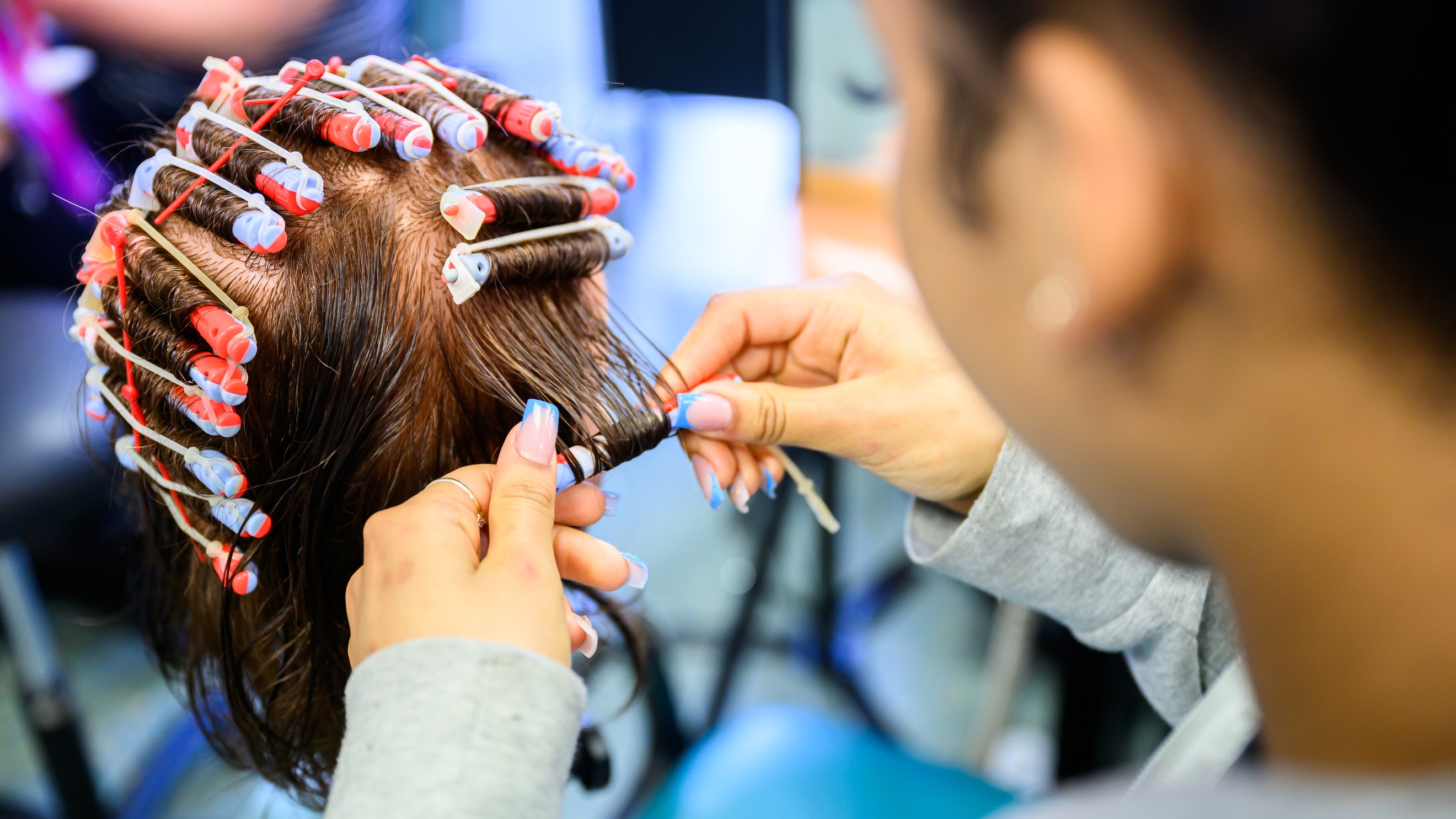 Ein Modepuppenkopf hat die Haare eingedreht. Neben dem Kopf steht eine Person, die eine Haarsträhne auf einen Lockenwickler aufdreht