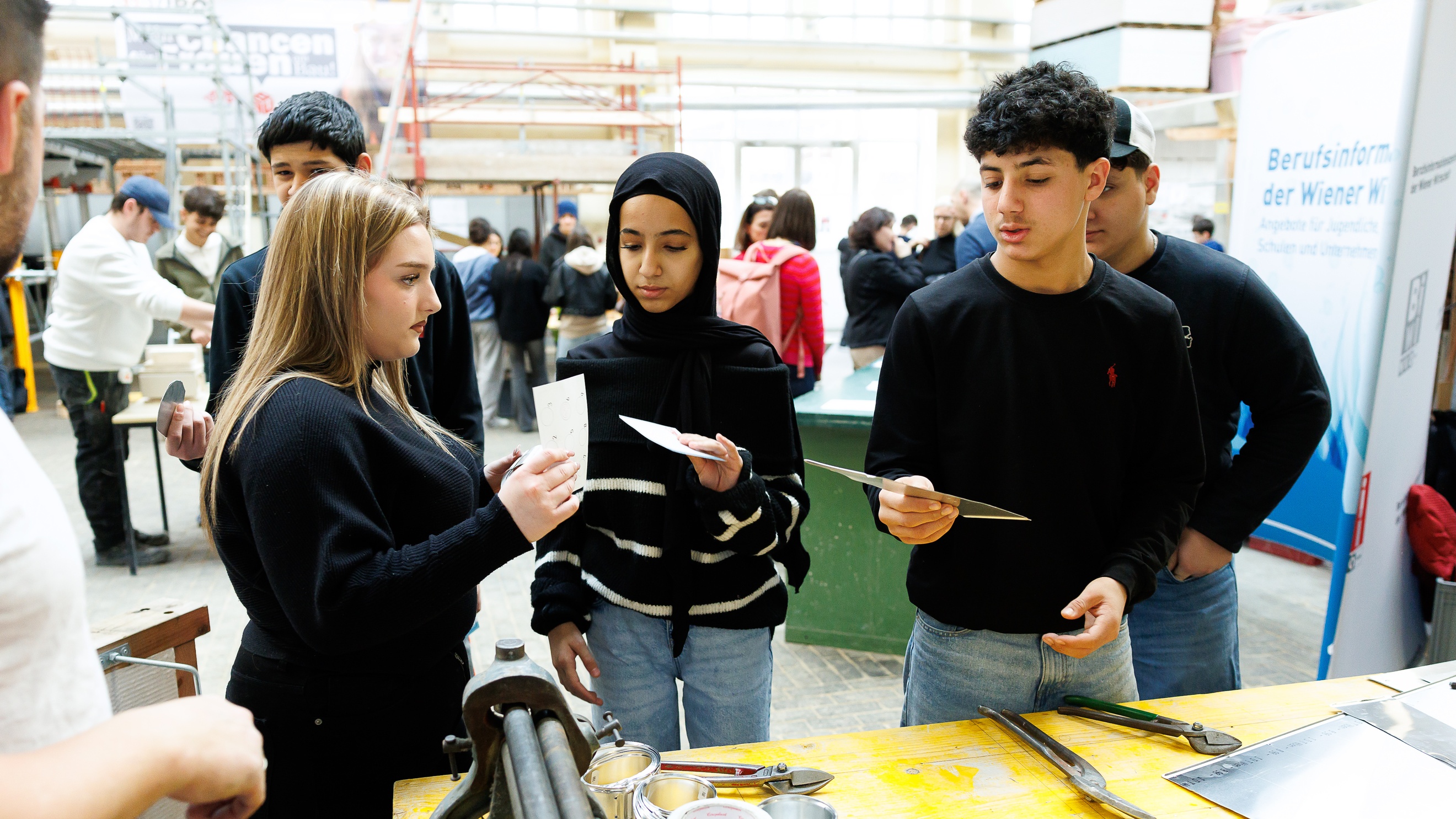 Fünf Jugendliche stehen auf einer Messe neben einem Tisch, auf dem verschiedene Werkzeuge zur Metallbearbeitung sind. Im Hintergrund sind weitere Personen, die hinter und neben Tischen stehen