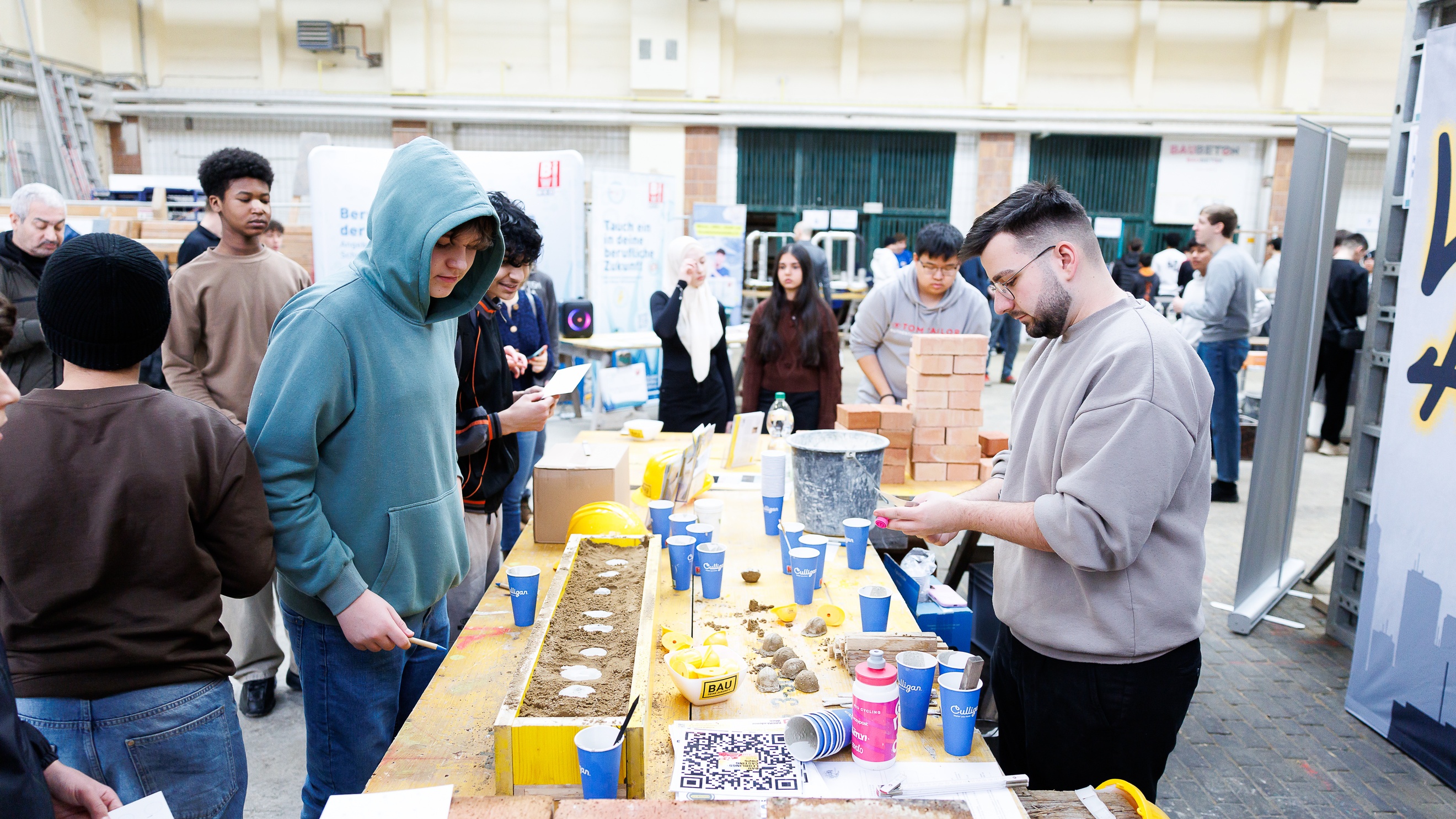 An einem langen Tisch stehen auf jeder Seite ein paar Personen. Auf dem Tisch sind Becher, Ziegel, ein Eimer, kleine Kugeln aus Sand, ein Bauhelm usw