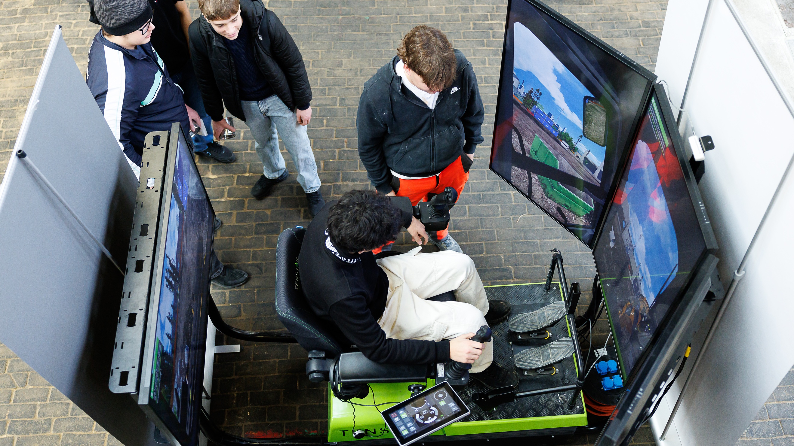 Eine jugendliche Person sitzt auf einem Simulator eines Gabelstaplers. Gegenüber der Person sind drei Bildschirme. Neben der Person stehen vier weitere Personen