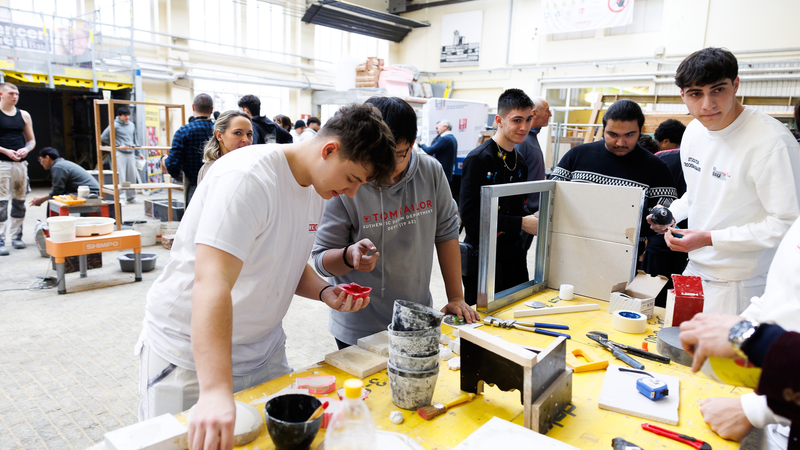 In einer großen Halle stehen um einen Tisch mehrere Personen. Auf dem Tisch sind verschiedene Werkzeuge wie Zangen, Maßbänder, Pinsel und weitere Utensilien