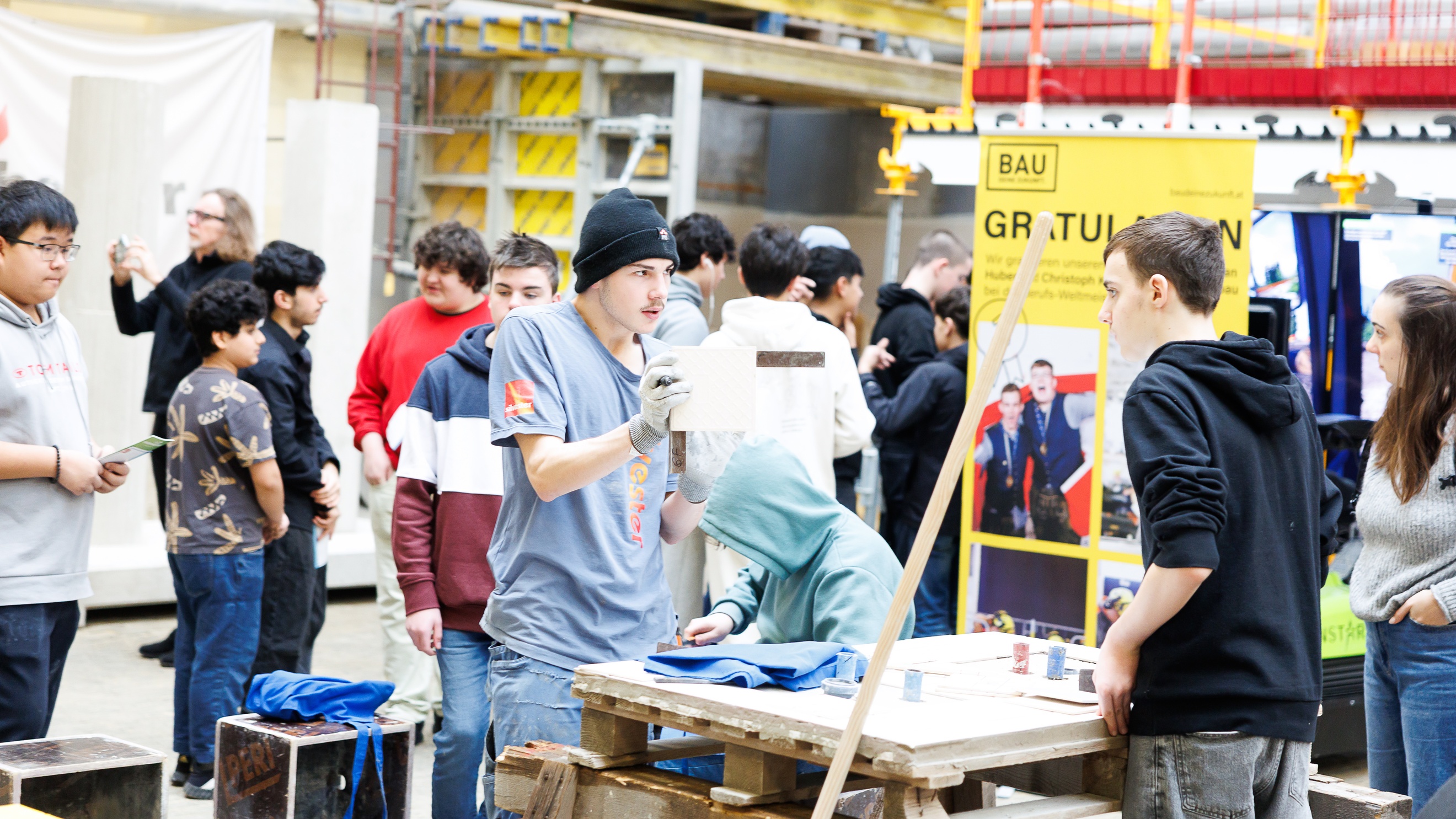 In einer großen Halle stehen sich zwei Personen an einem Pallettentisch gegenüber. Die linke Person zeigt der anderen Person eine Fliese, an die sie ein Lineal hält. Im Hintergrund sind viele weitere Personen