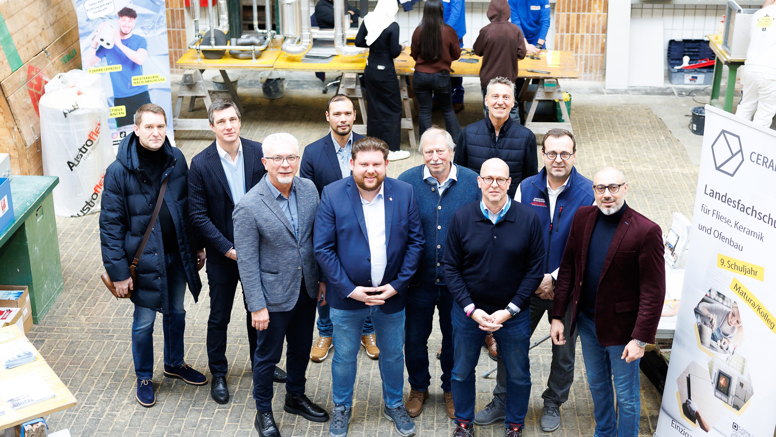 Porträt von zehn Personen, die nebeneinander in einer Halle stehen. Neben ihnen ist ein Banner, auf dem Landesfachschule für Fliese Keramik und Ofenbau steht. Im Hintergrund stehen an einem langen Tisch drei Personen
