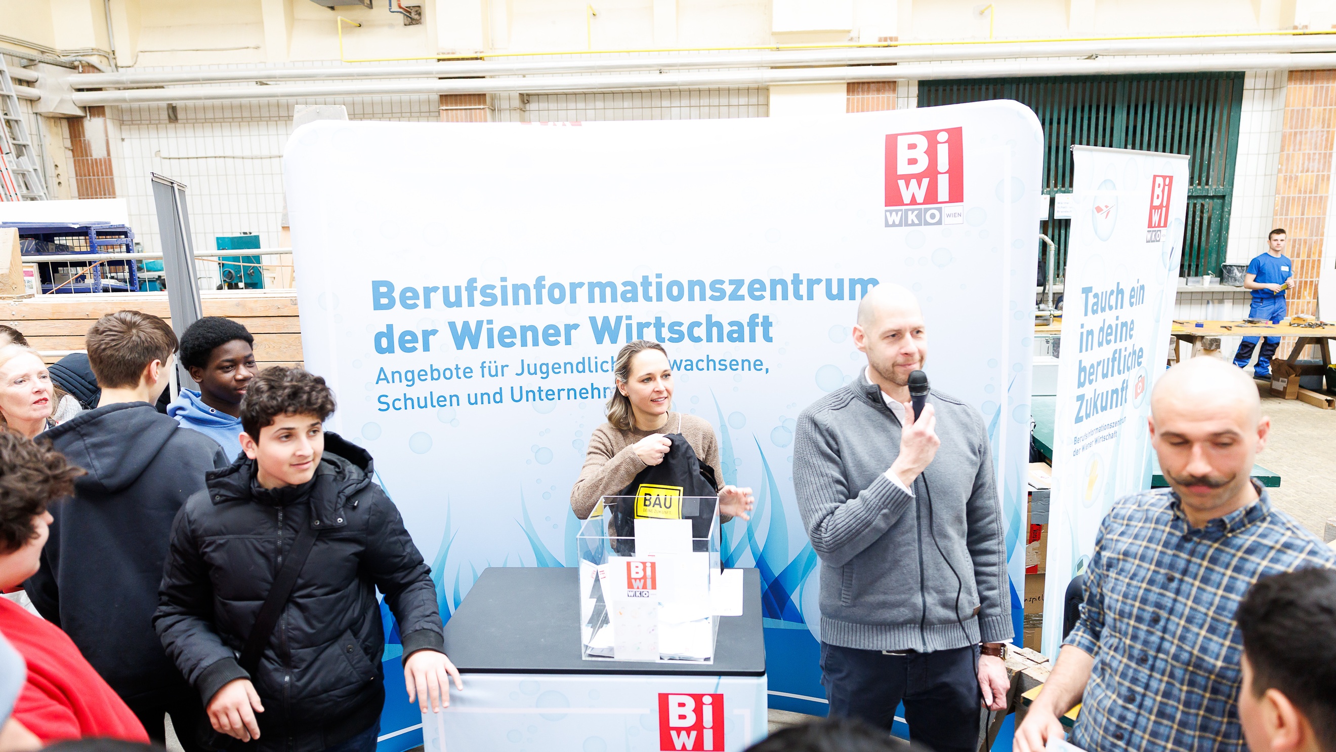 An einem hohen Tisch stehen zwei Personen. Die rechte Person hält ein Mikrofon vor den Mund. Hinter ihnen ist ein großer Banner, auf dem Berufsinformationszentrum der Wiener Wirtschaft steht. Um den Tisch stehen weitere jugendliche und erwachsene Personen