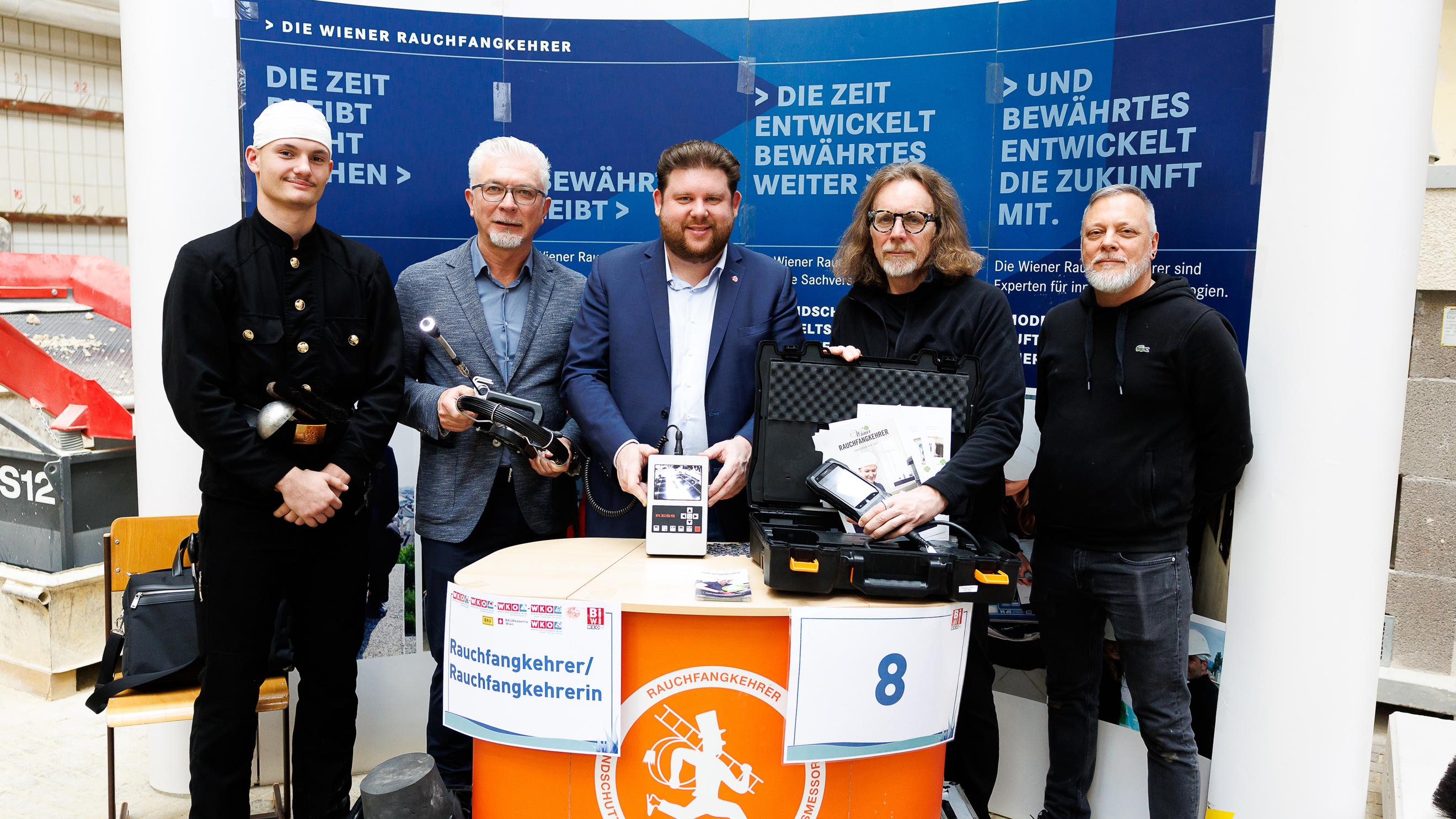 Porträt von fünf Personen, die hinter einem Tisch stehen. Am Tisch hängt ein Zettel, auf dem Rauchfangkehrerin steht. Eine Person trägt die Arbeitskleidung eines Rauchfangkehrers