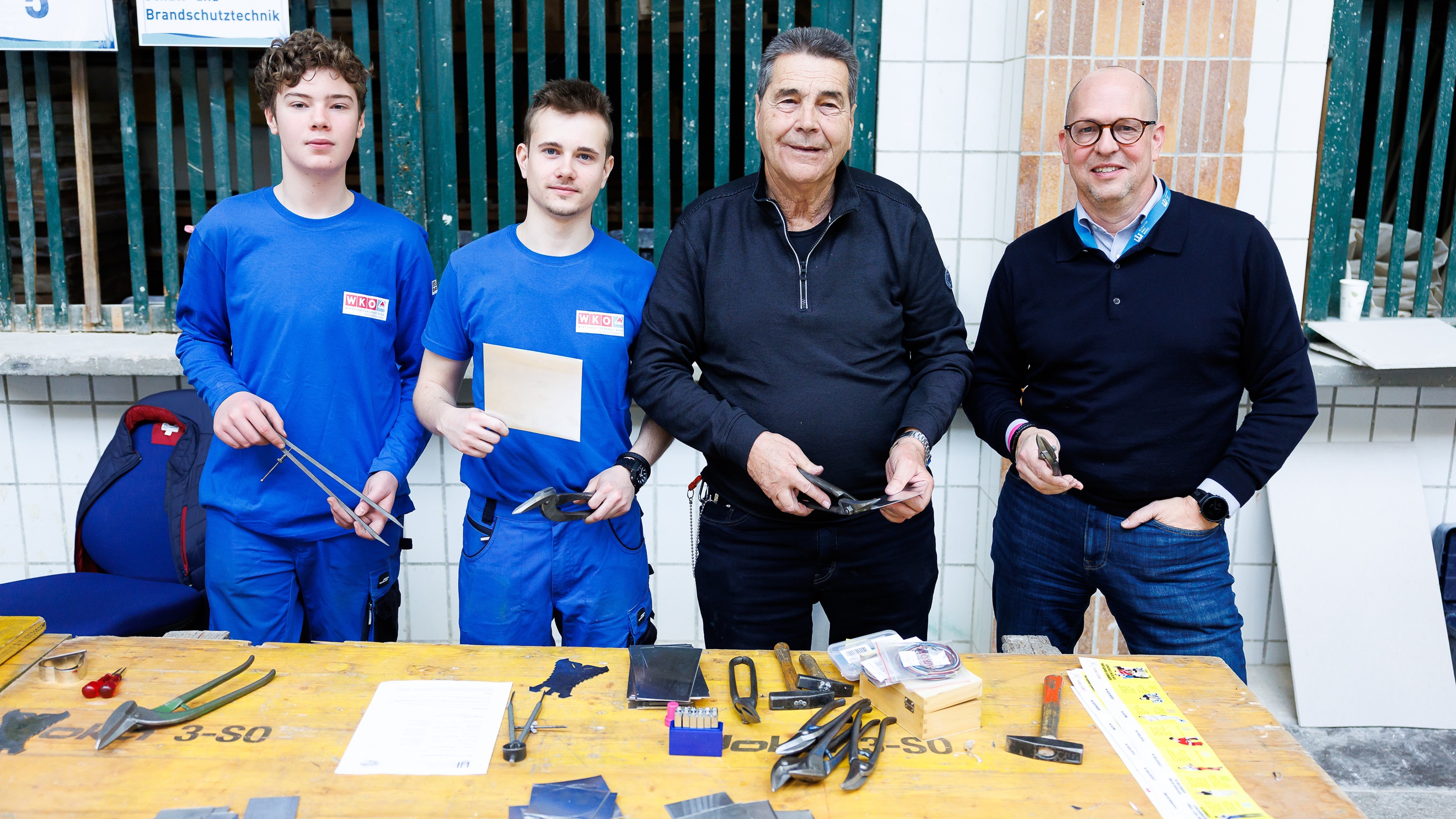 Porträt von zwei jugendlichen und zwei erwachsenen Personen, die hinter einem Tisch stehen. Auf dem Tisch liegen verschiedene Werkzeuge. Hinter ihnen hängt ein Zettel, auf dem Schall- und Brandschutztechnik steht