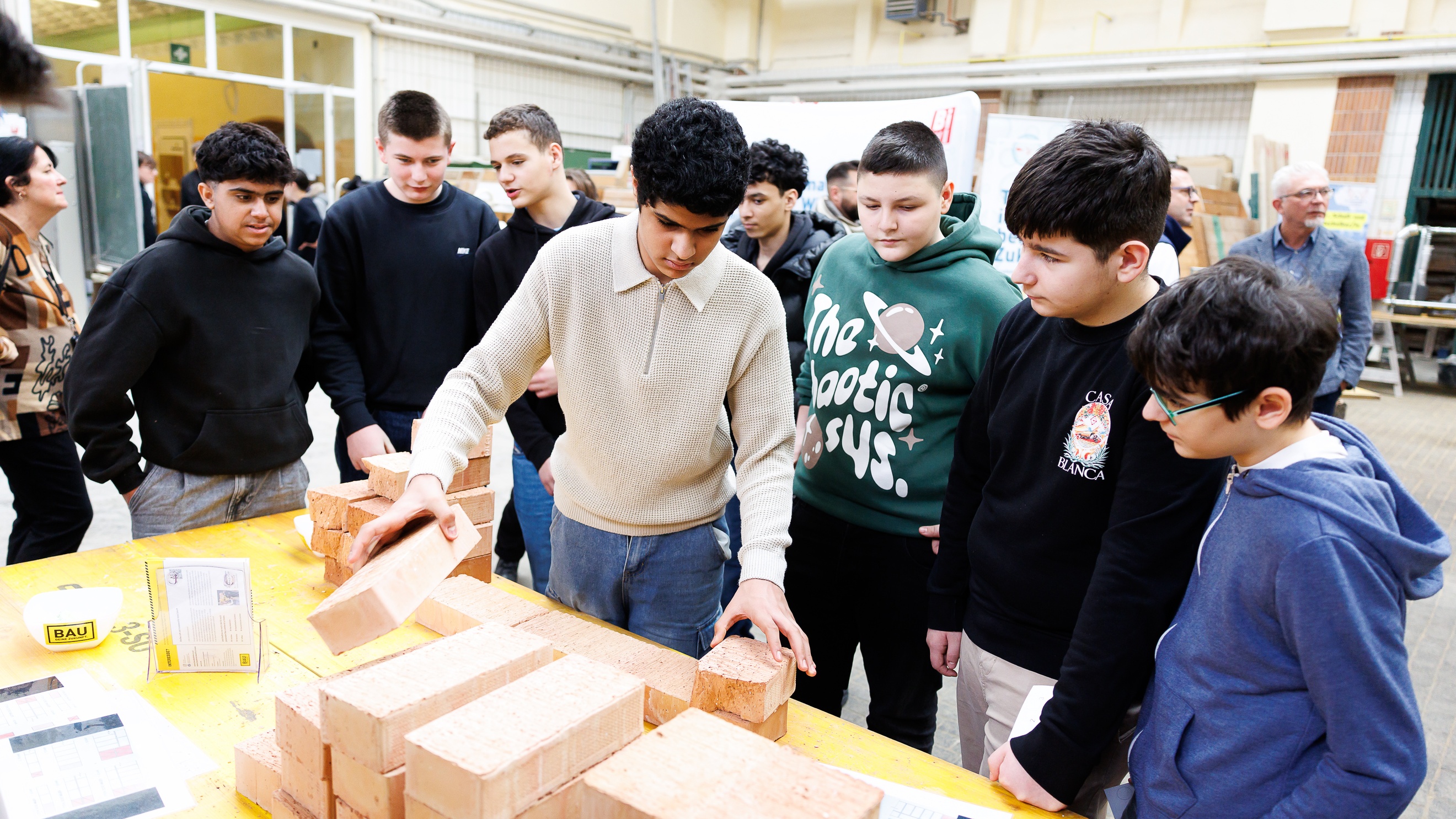 An einem Tisch stehen mehrere jugendliche Personen. Auf dem Tisch liegen Ziegelsteine