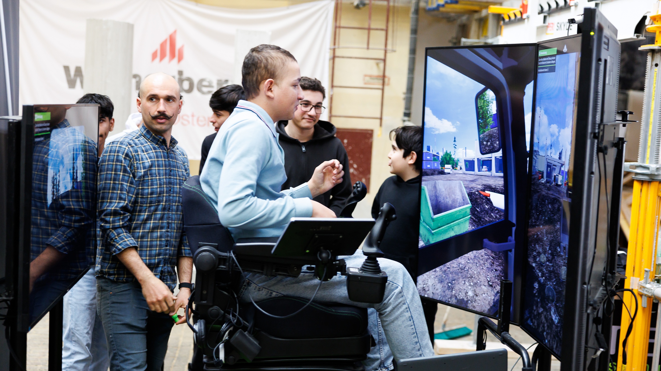 Seitliche Aufnahme einer jugendlichen Person in einem Sessel, der links und rechts einen Steuerknüppel montiert hat. Vor der Person steht ein Bildschirm, auf dem ein Bagger ein Loch gräbt. Neben der Person stehen eine erwachsene und eine jugendliche Personen, die auf den Bildschirm blicken