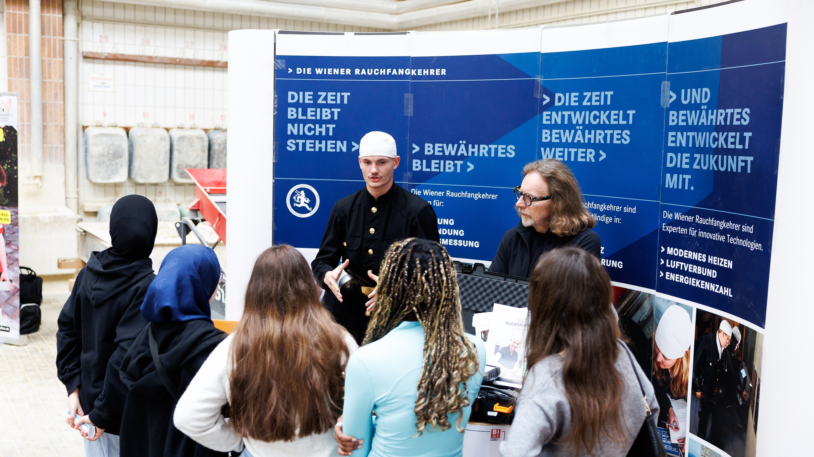 Zwei Personen stehen an einem Tisch nebeneinander. Hinter ihnen ist ein großer Banner, auf dem die Wiener Rauchfangkehrer steht. Die linke Person trägt ein Rauchfangkehreroutfit. Zu dieser Person blicken fünf jugendliche Personen
