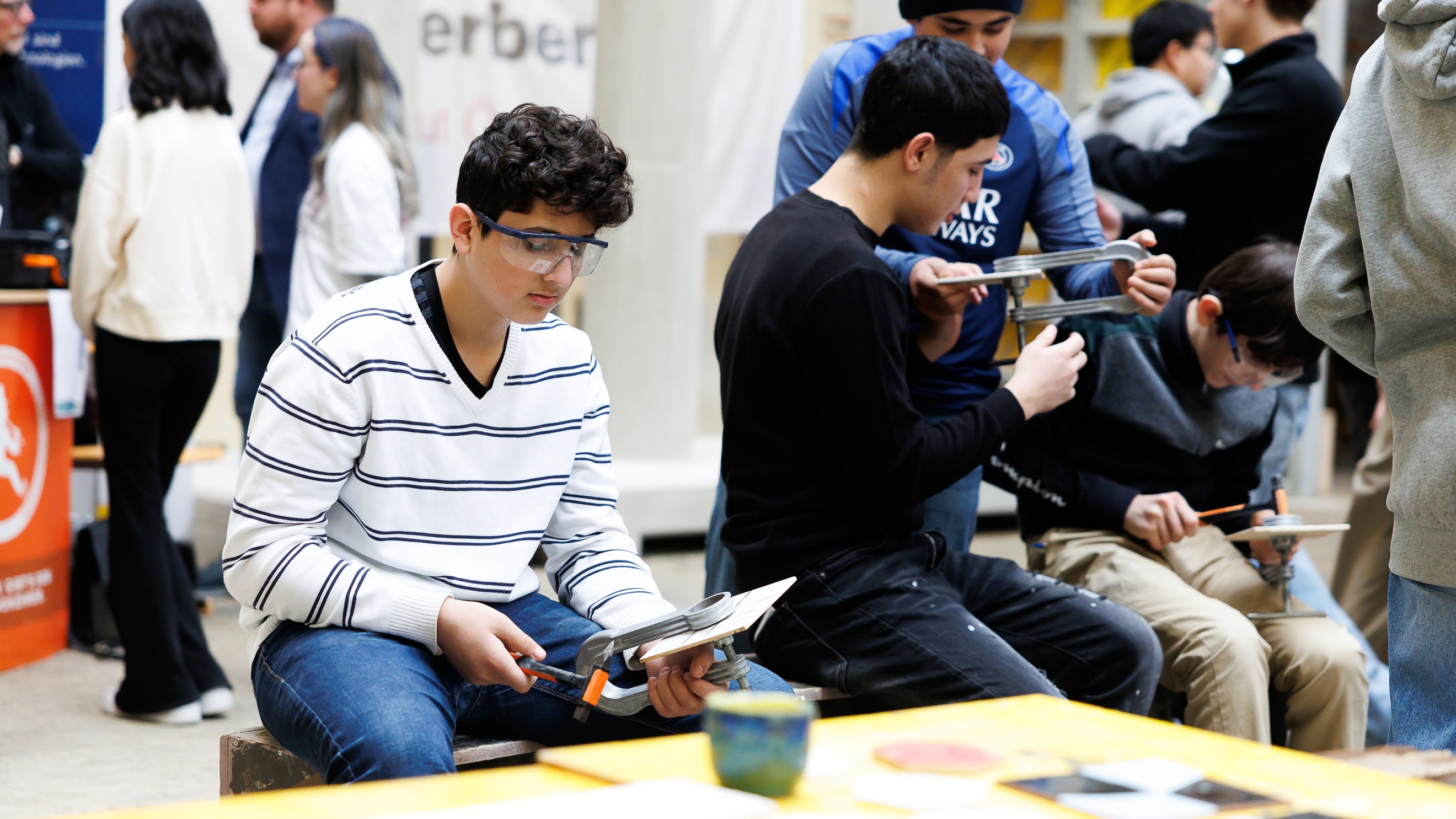 Frontale Aufnahme einer jugendlichen Person mit Schutzbrille, die eine Zwinge hält, in der etwas eingespannt ist. Daneben sitzen weitere jugendliche Personen mit Zwingen
