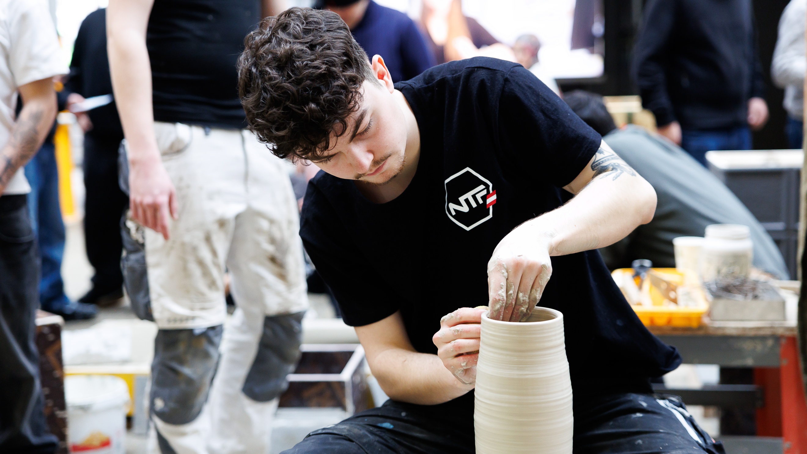 Frontale Aufnahme einer Person, die ein Objekt töpfert. Im Hintergrund sind weitere Personen