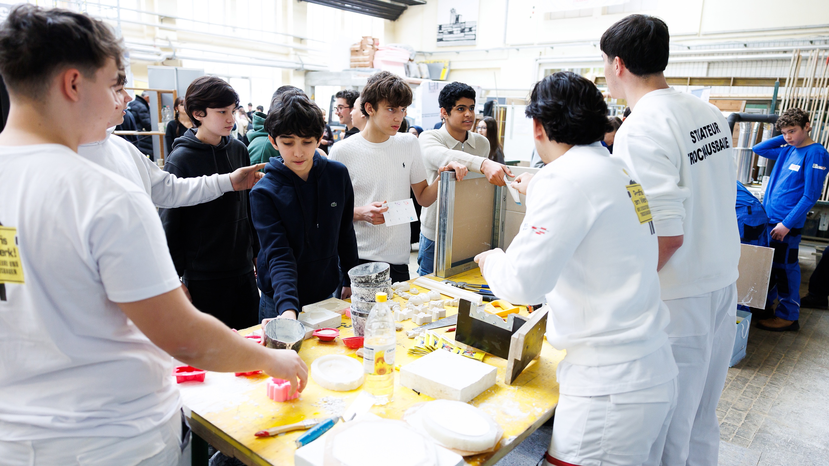 In einer großen Halle stehen um einen Tisch mehrere Personen. Auf dem Tisch sind verschiedene Werkzeuge wie Zangen, Maßbänder, Pinsel und weitere Utensilien