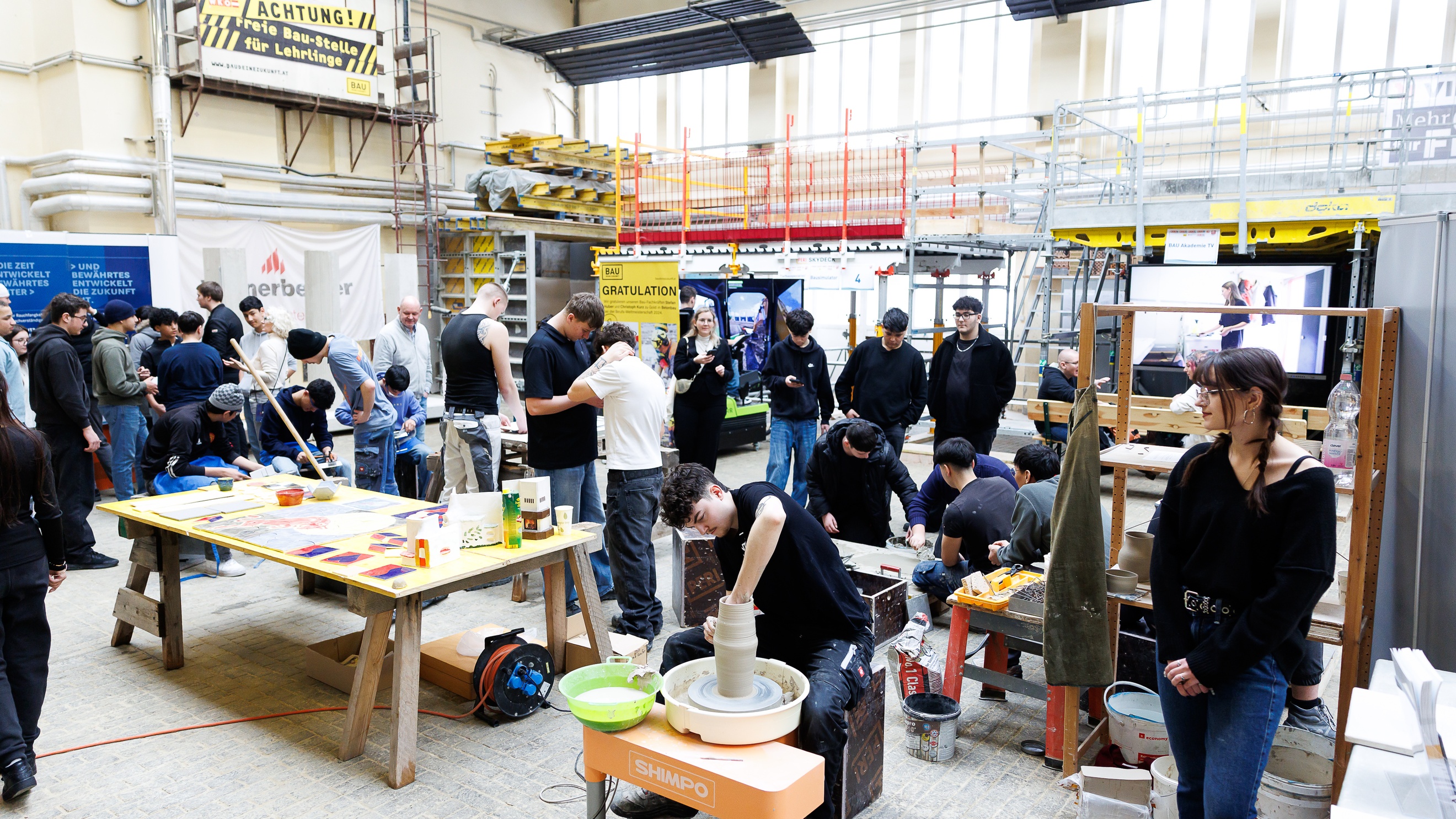 Aufnahme einer großen Halle, in der an verschiedenen Stationen erwachsene Personen und Jugendliche stehen. An einer Station töpfert eine Person etwas. An einer anderen Station rührt eine Person etwas in einem Eimer um