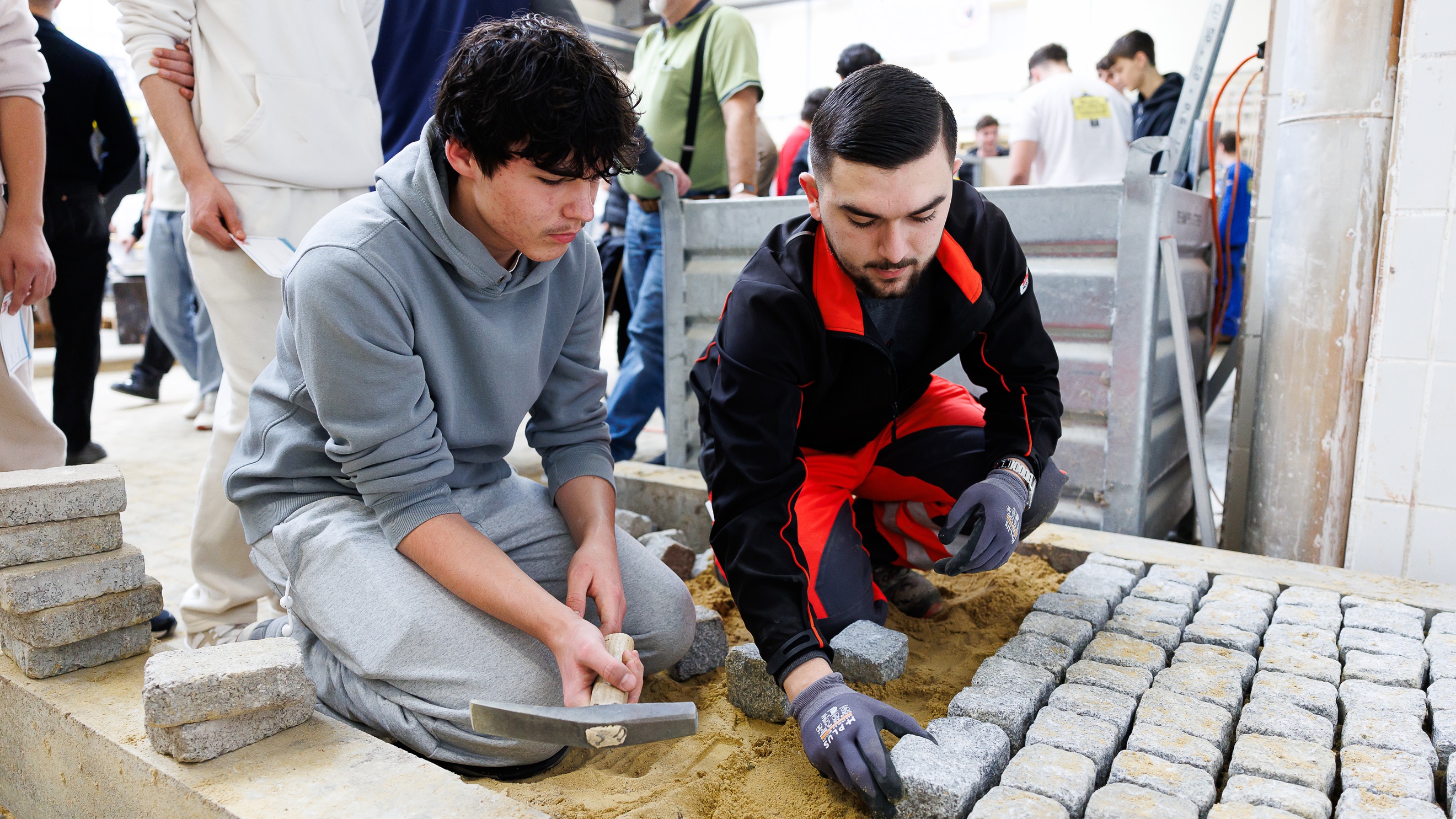 In einem kleinen Bereich, der von niedrigen Betonklötzen begrenzt wird, hocken nebeneinander zwei Personen. Die rechte Person legt kleine Pflastersteine auf den Bereich, der mit Sand ausgelegt ist. Die linke Person hält eine Spitzhacke