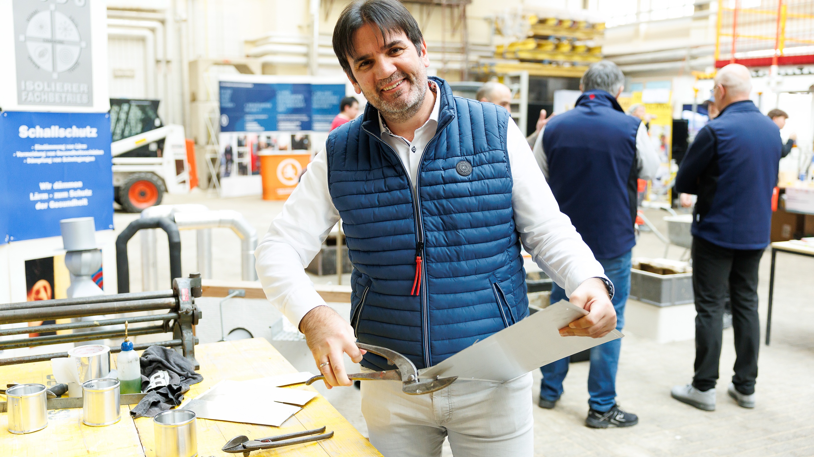 Frontale Aufnahme einer Person, die neben einem Tisch steht, ein dünnes Metallbrett hält und mit einer Metallschere in das Metall schneidet