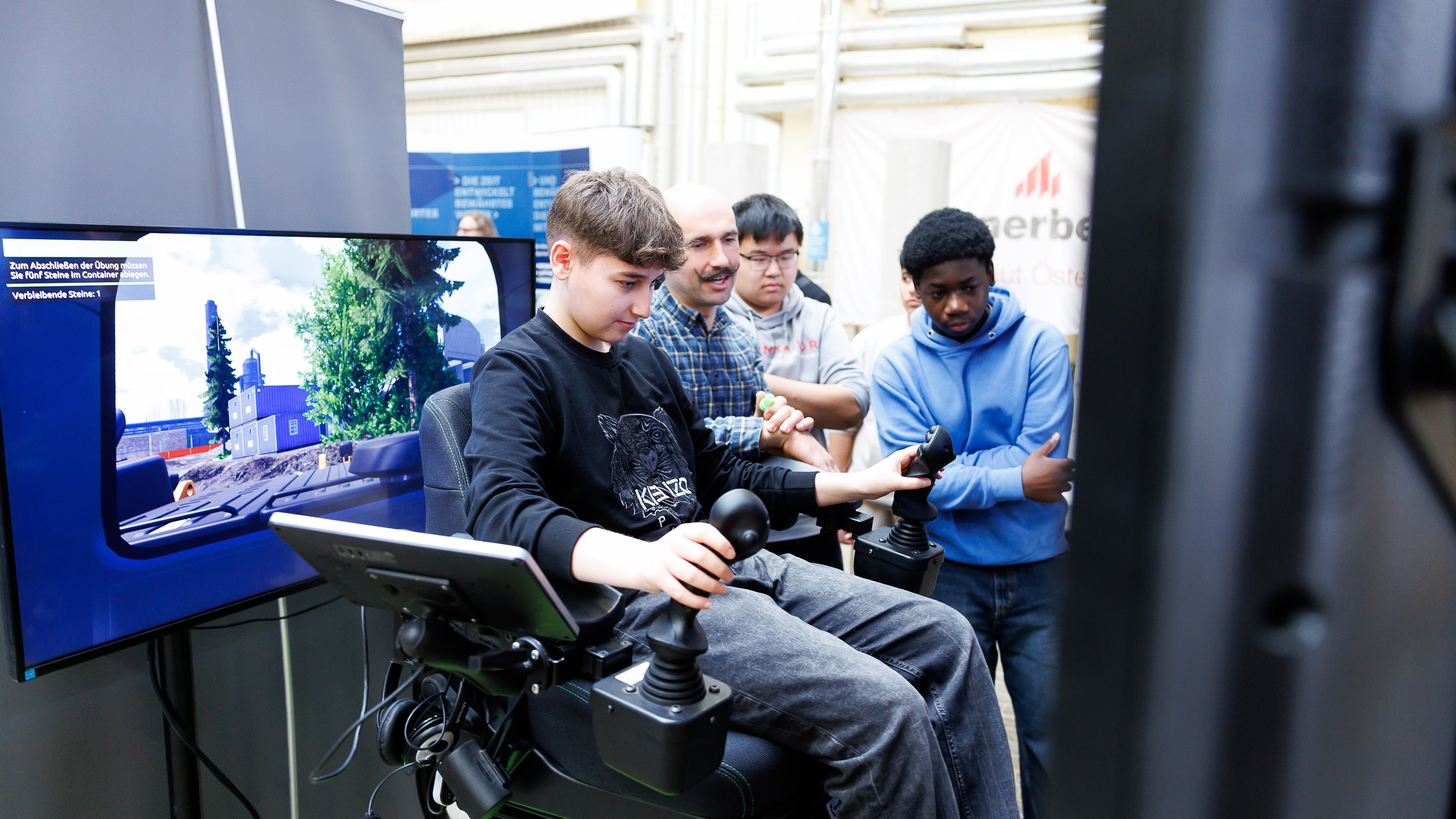 Eine jugendliche Person sitzt in einem Sessel, der links und rechts einen Steuerknüppel montiert hat. Vor der Person steht ein Bildschirm. Neben der Person stehen eine erwachsene und zwei jugendliche Personen, die auf den Bildschirm blicken