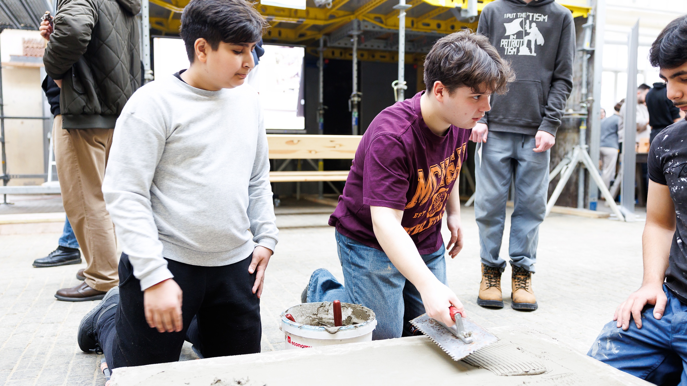 Drei jugendliche Personen knien neben einer Betonplatte, die am Boden eines großen Raums liegt. Die mittlere Person verteilt Beton auf der Platte. Die anderen sehen zu