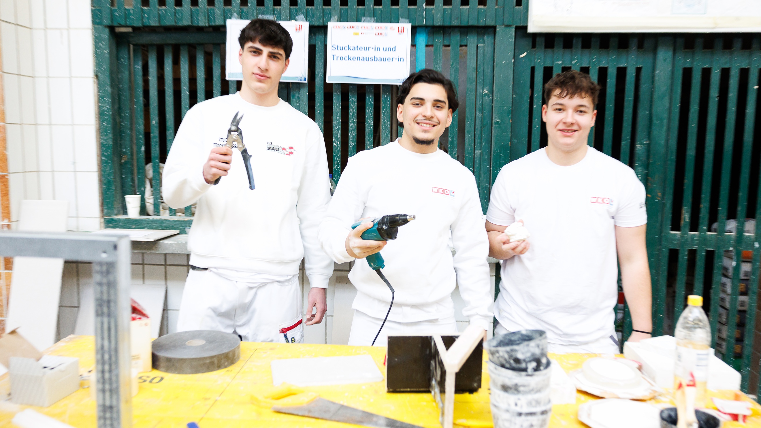 Porträt von drei Personen in hellen Oberteilen, die nebeneinander hinter einem Tisch stehen und jeweils ein unterschiedliches Werkzeug halten. Hinter ihnen hängt ein Zettel, auf dem Stuckateurin und Trockenausbauerin steht