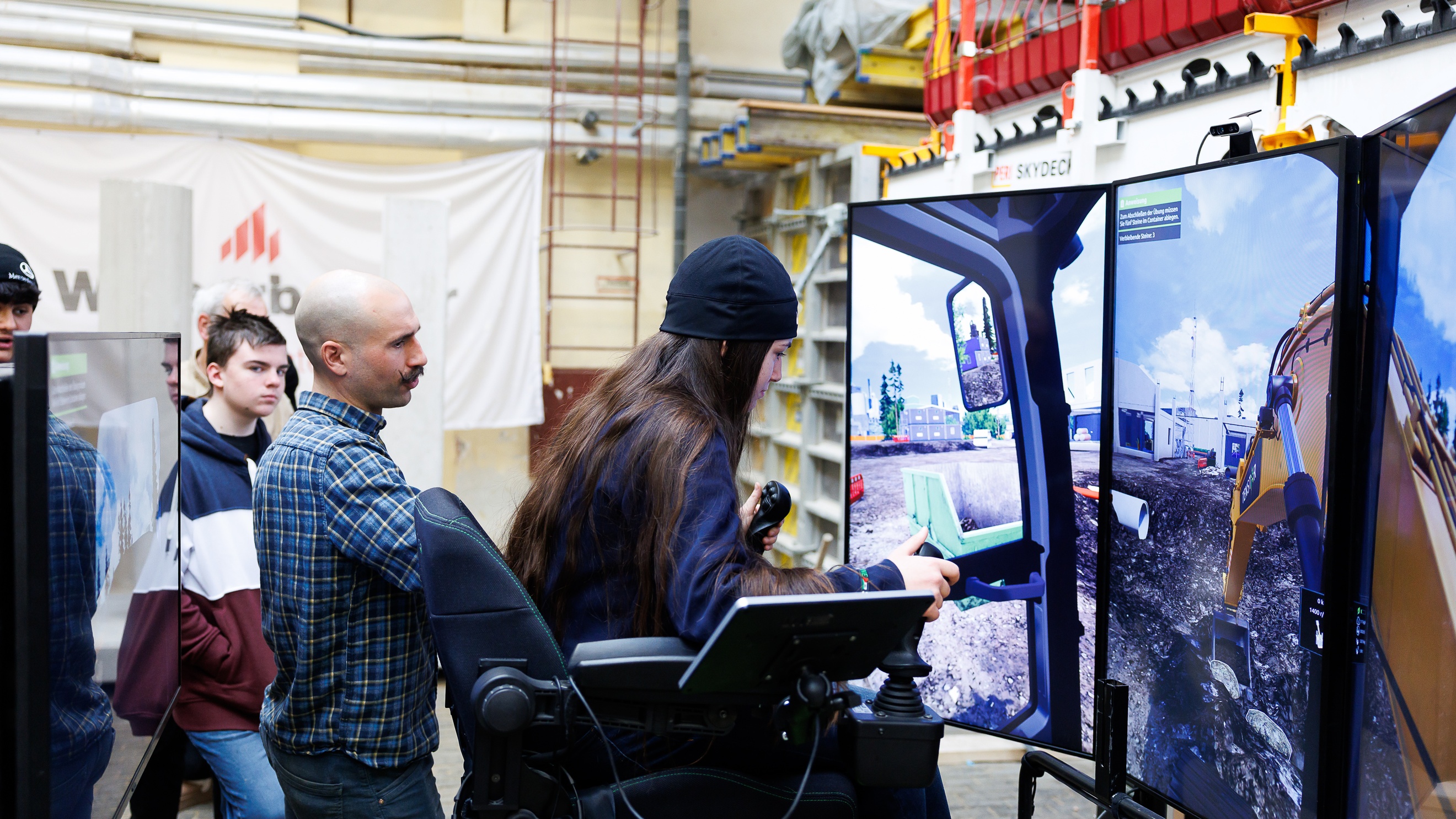 Seitliche Aufnahme einer jugendlichen Person in einem Sessel, der links und rechts einen Steuerknüppel montiert hat. Vor der Person steht ein Bildschirm, auf dem ein Bagger ein Loch gräbt. Neben der Person stehen eine erwachsene und eine jugendliche Personen, die auf den Bildschirm blicken