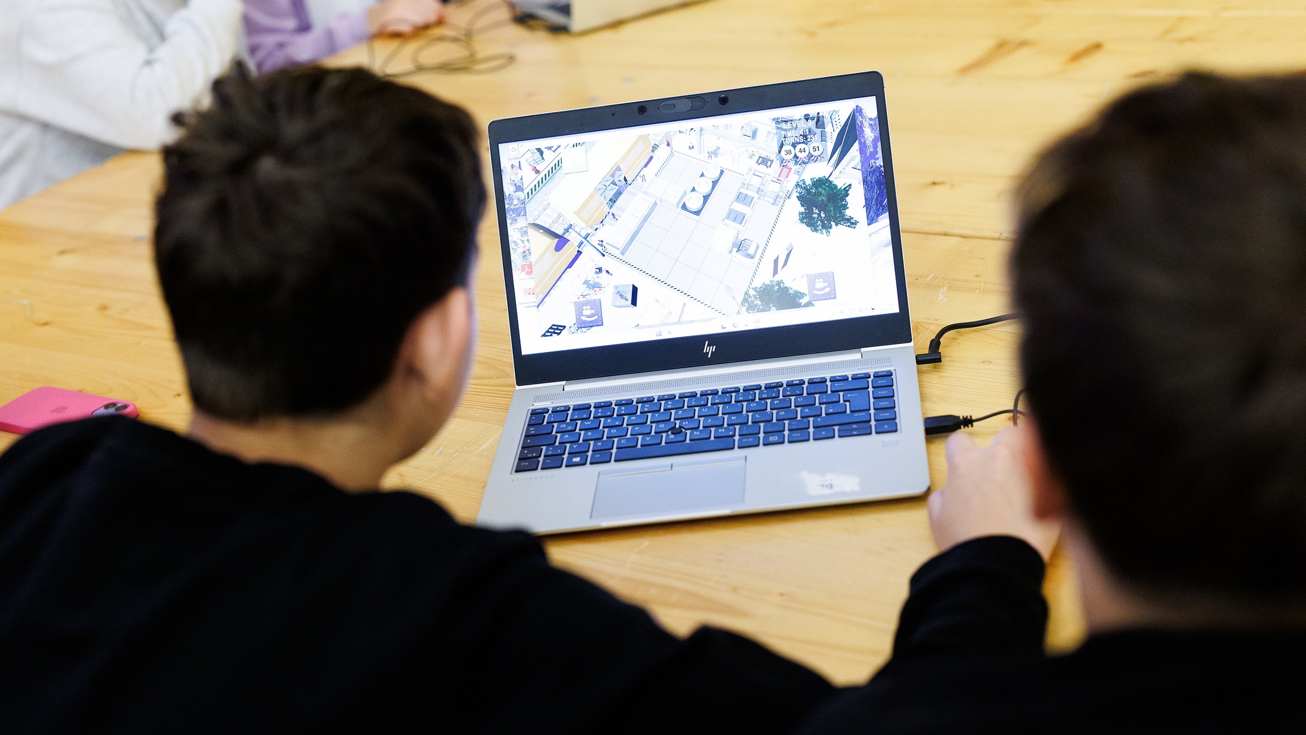 Rückenansicht zweier Personen, die nebeneinander an einem Holztisch sitzen und gemeinsam auf den Bildschirm eines Laptops blicken. Auf dem Bildschirm ist ein Warenlager