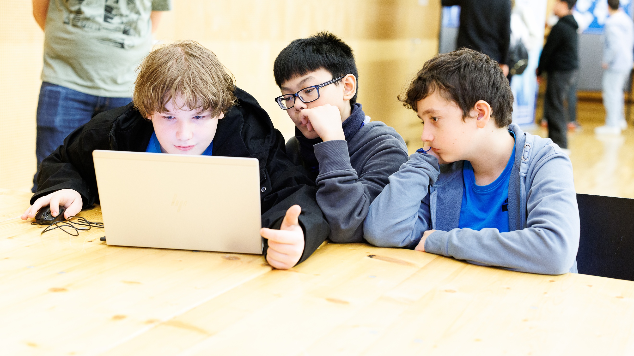Drei Kinder sitzen nebeneinander an einem Holztisch und blicken auf den Bildschirm eines Laptops. Daneben steht eine Person. Im Hintergrund sind weitere Personen