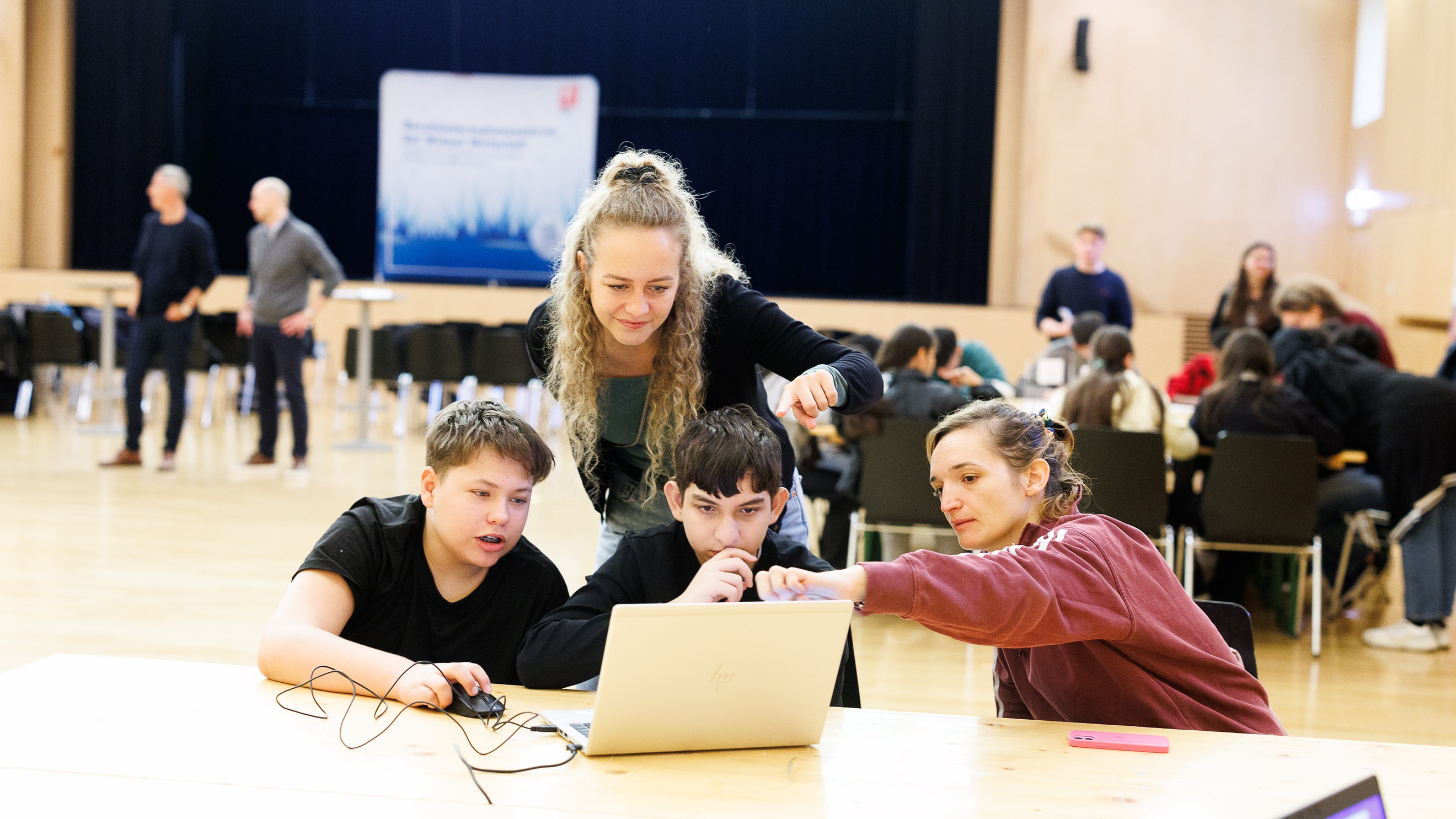 Zwei Kinder sitzen nebeneinander an einem Holztisch und blicken auf den Bildschirm eines Laptops. Neben ihnen sitzt eine Person, die auf den Laptop zeigt. Daneben steht eine weitere Person. Im Hintergrund sind weitere Personen
