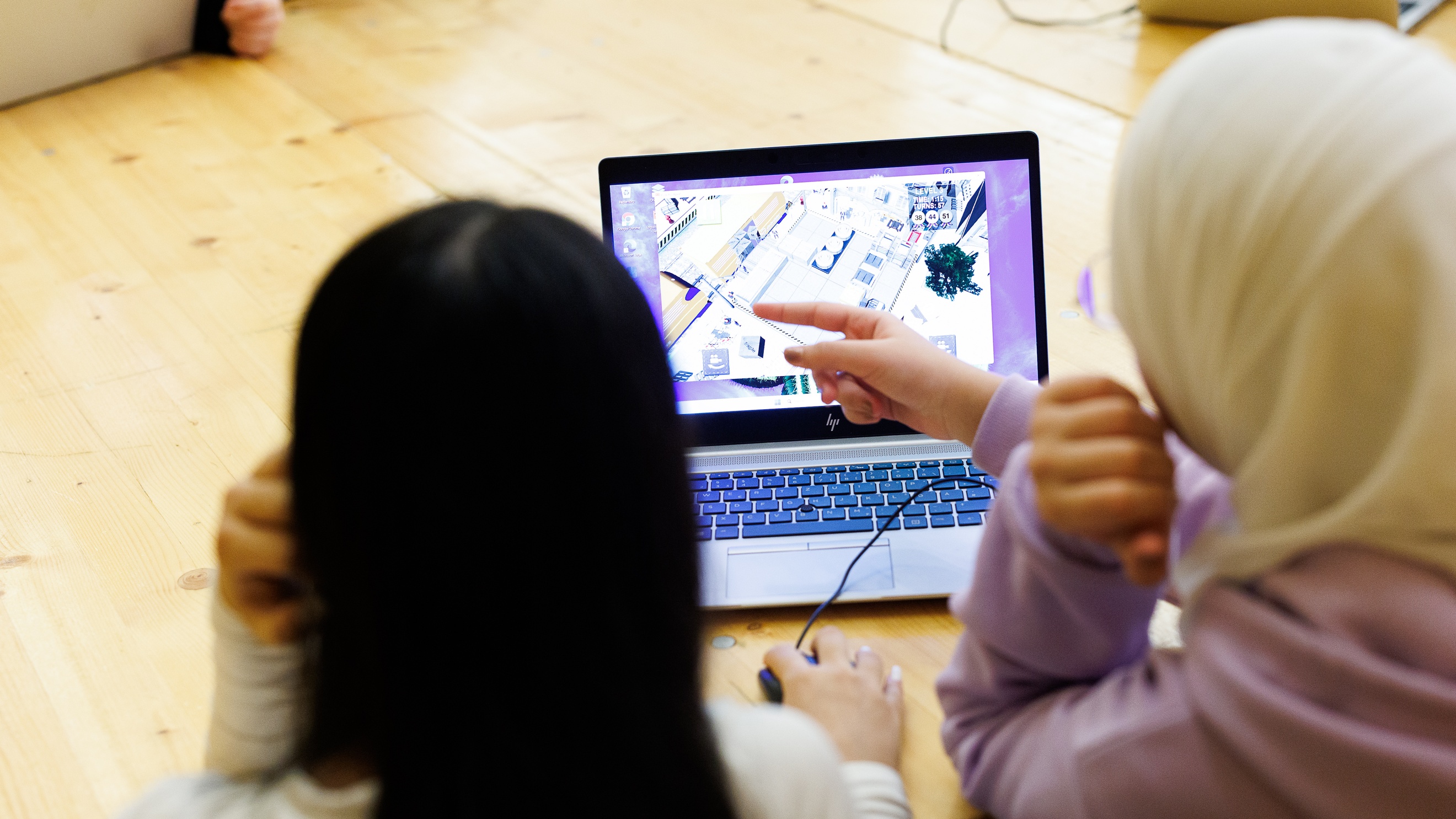 Rückenansicht zweier Personen, die nebeneinander an einem Holztisch sitzen und gemeinsam auf den Bildschirm eines Laptops blicken. Auf dem Bildschirm ist ein Warenlager