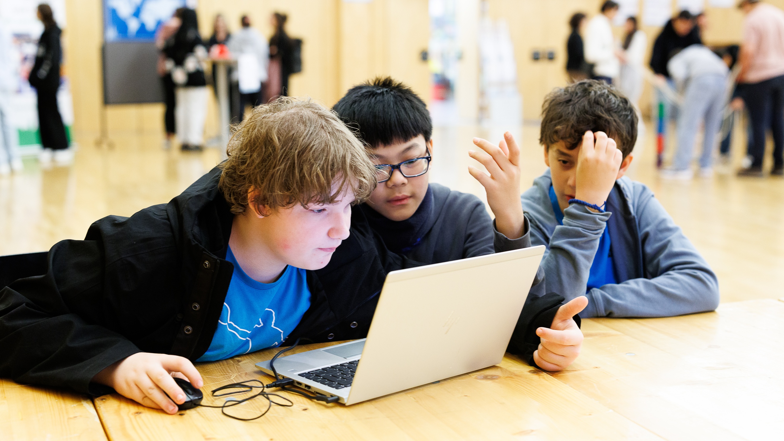 rei Kinder sitzen nebeneinander an einem Holztisch und blicken auf den Bildschirm eines Laptops. Im Hintergrund sind weitere Personen