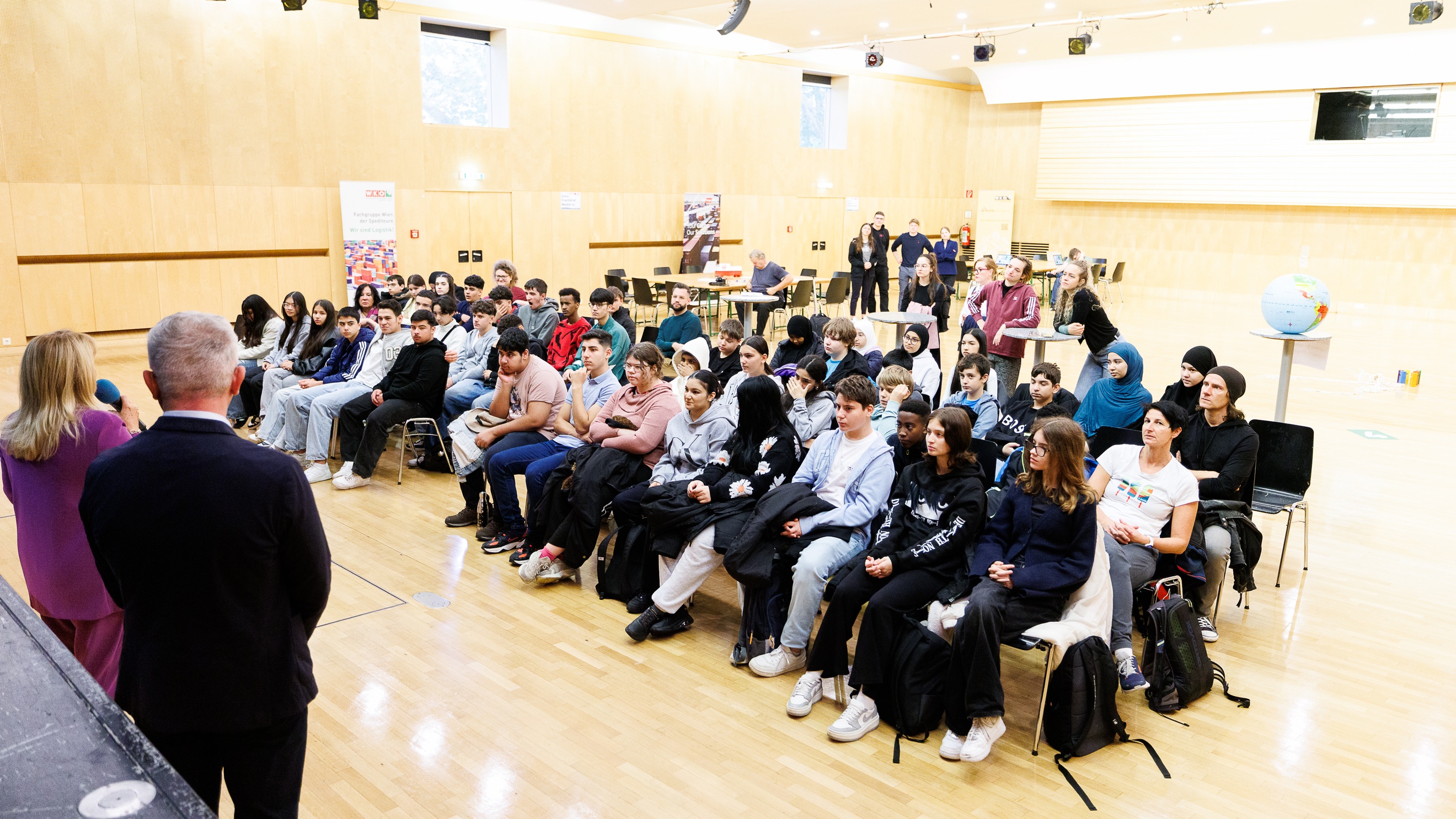 In einem großen Saal mit Holzboden und Holzvertäfelungen an der Wand sitzen in einem Publikum viele Jugendliche. Ihnen gegenüber stehen zwei Personen in Sakkos. Die linke Person spricht in ein Mikrofon. Auf einem Stelltisch im Hintergrund liegt ein Globus