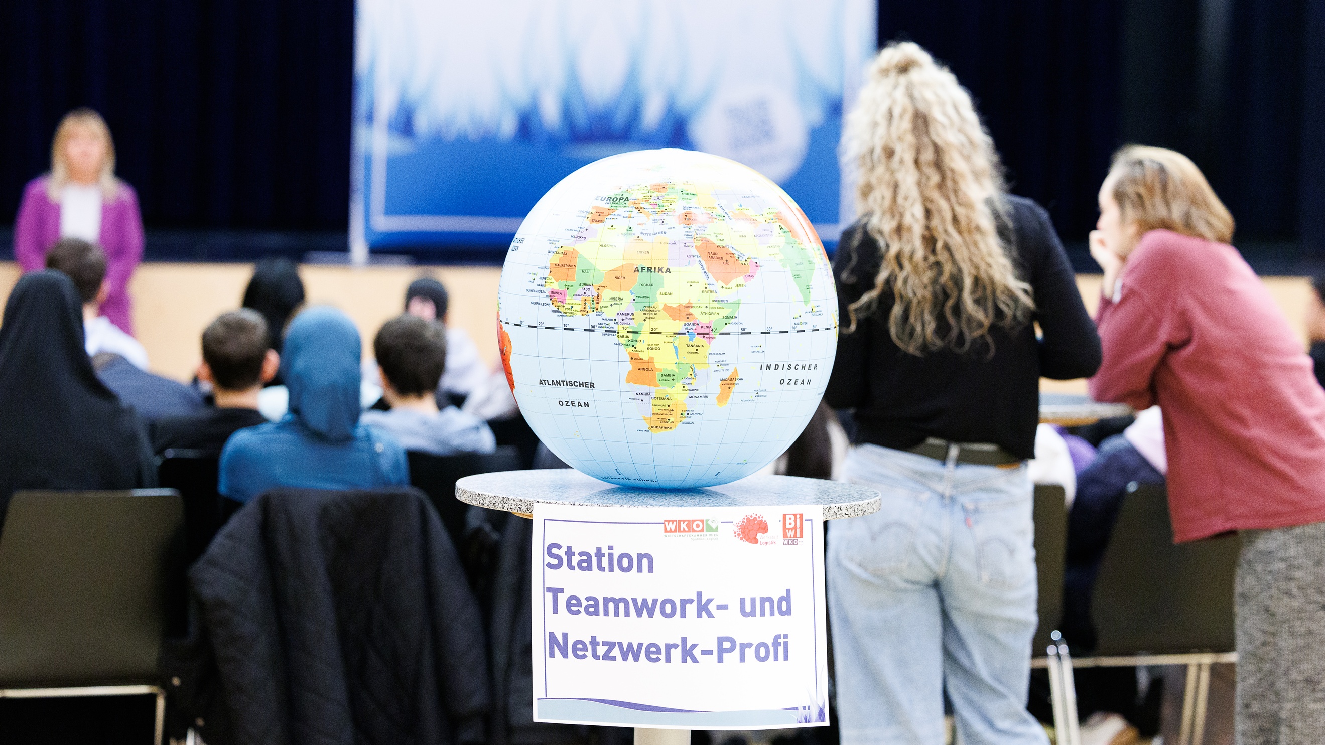 Auf einem Stelltisch liegt ein Globus. Am Stelltisch ist ein Zettel angeklebt, auf dem Station Teamwork- und Netzwerk-Profi steht. Im Hintergrund ist die Rückenansicht eines sitzenden Publikums. Dem Publikum gegenüber steht eine Person