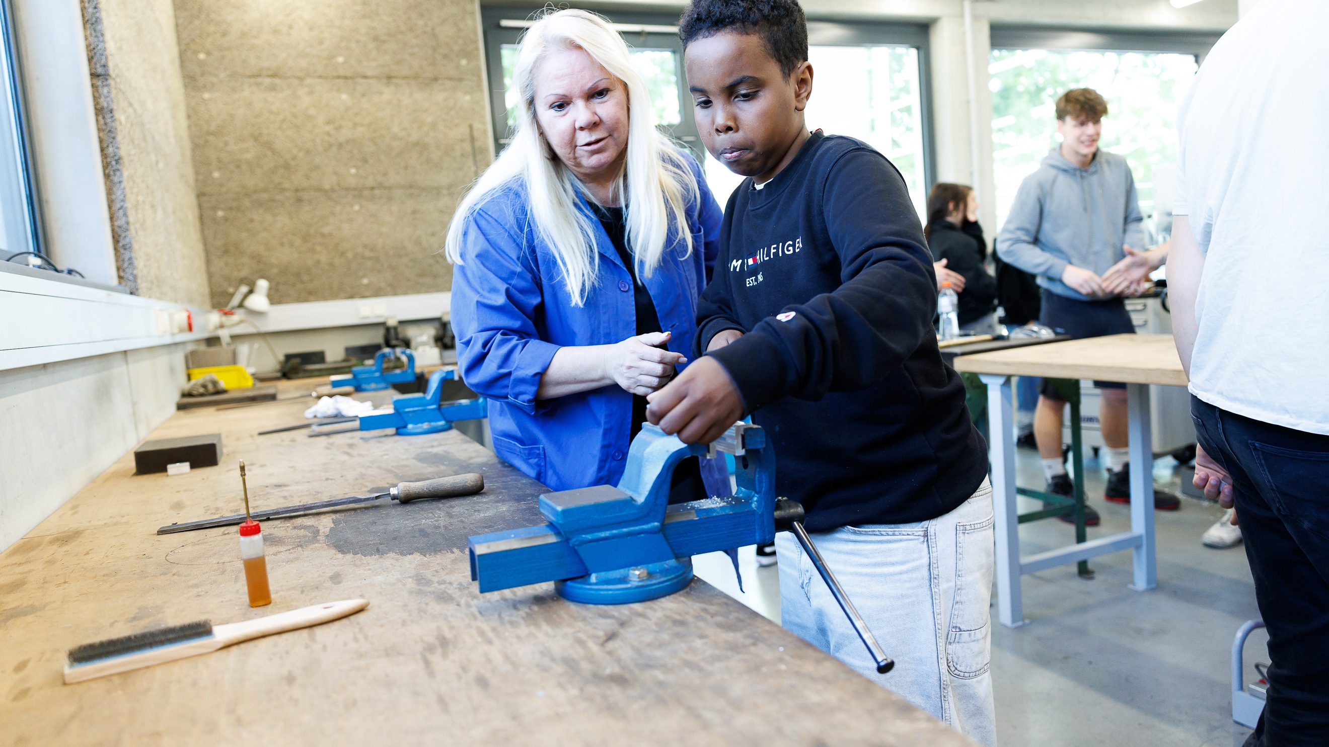 Eine erwachsene und eine jugendliche Person stehen nebeneinander an einem Tisch in einer Werkstatt. Auf dem Tisch liegen verschiedene Werkzeuge. Die erwachsene Person erklärt etwas