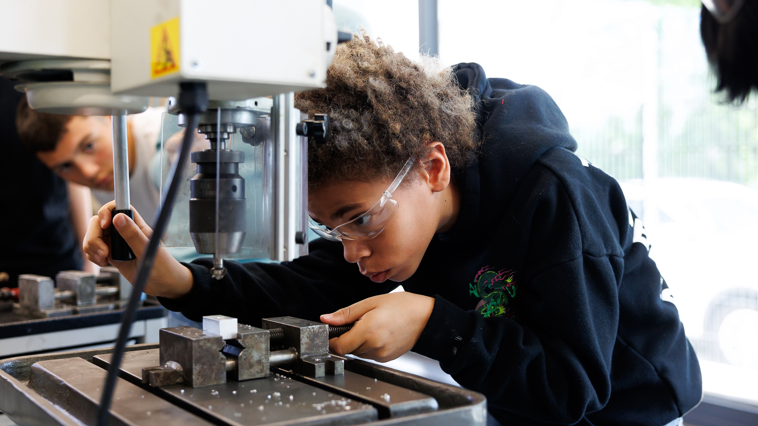 Ein Kind mit Schutzbrille bedient den Hebel eines Metallbohrers. In der Bohrmaschine ist ein kleines Objekt eingespannt