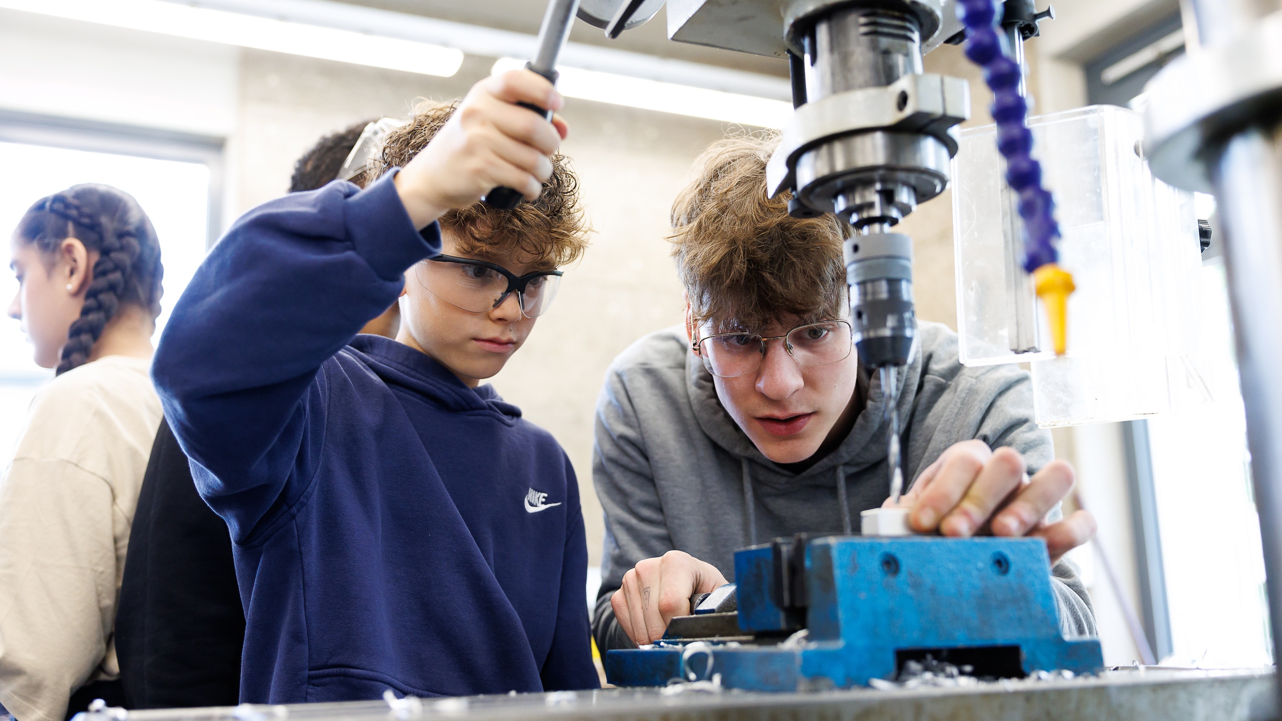 Ein Kind mit Schutzbrille bedient den Hebel eines Metallbohrers. Neben ihr steht eine weitere Person, die ein Objekt unter dem Bohrer mit einer Hand festhält und darauf blickt