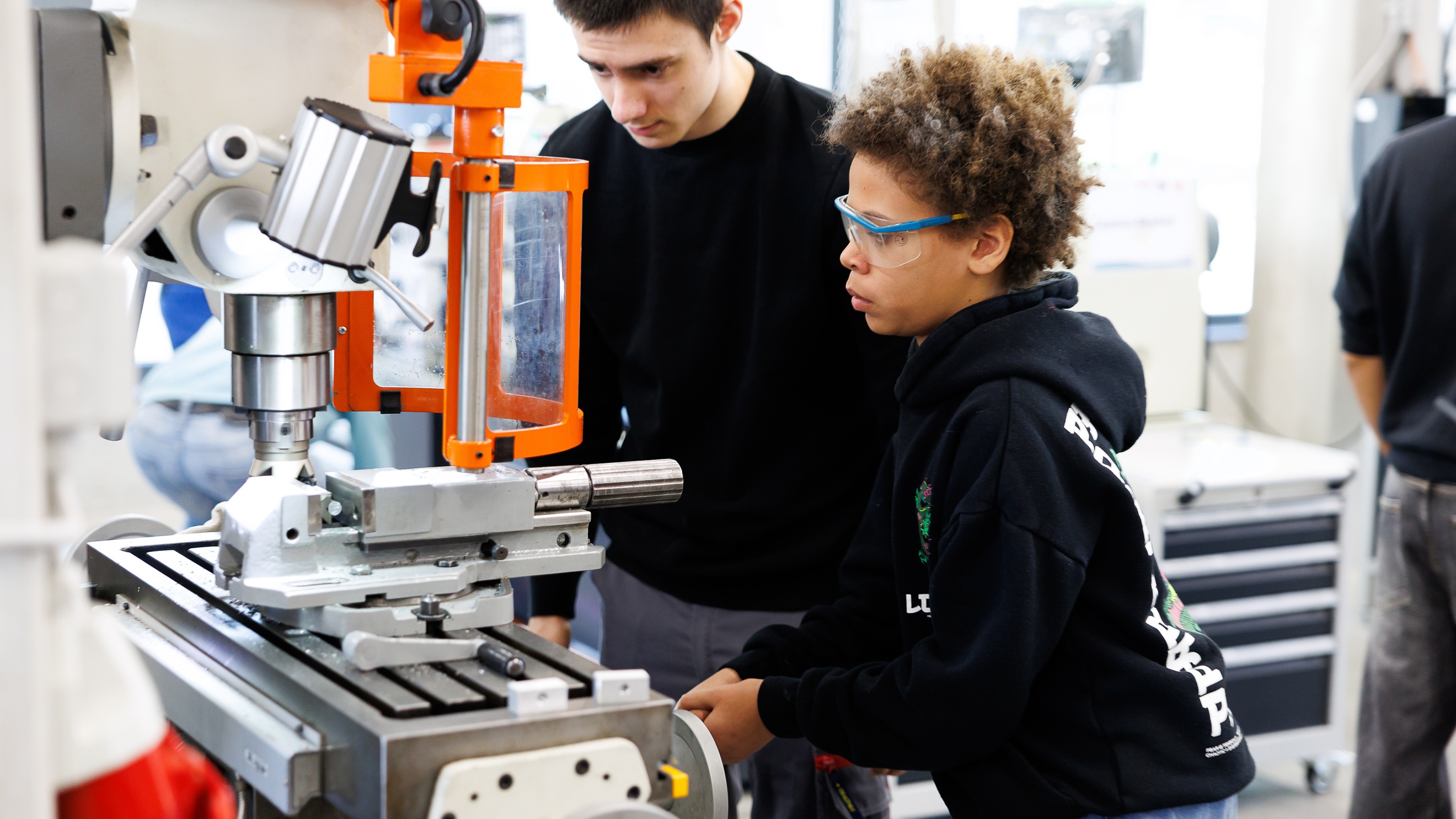 Ein Kind mit Schutzbrille bedient ein Gerät in einer Werkstatt mit einer Kurbel, die an der Seite des Geräts befestigt ist. Neben ihr steht eine weitere Person