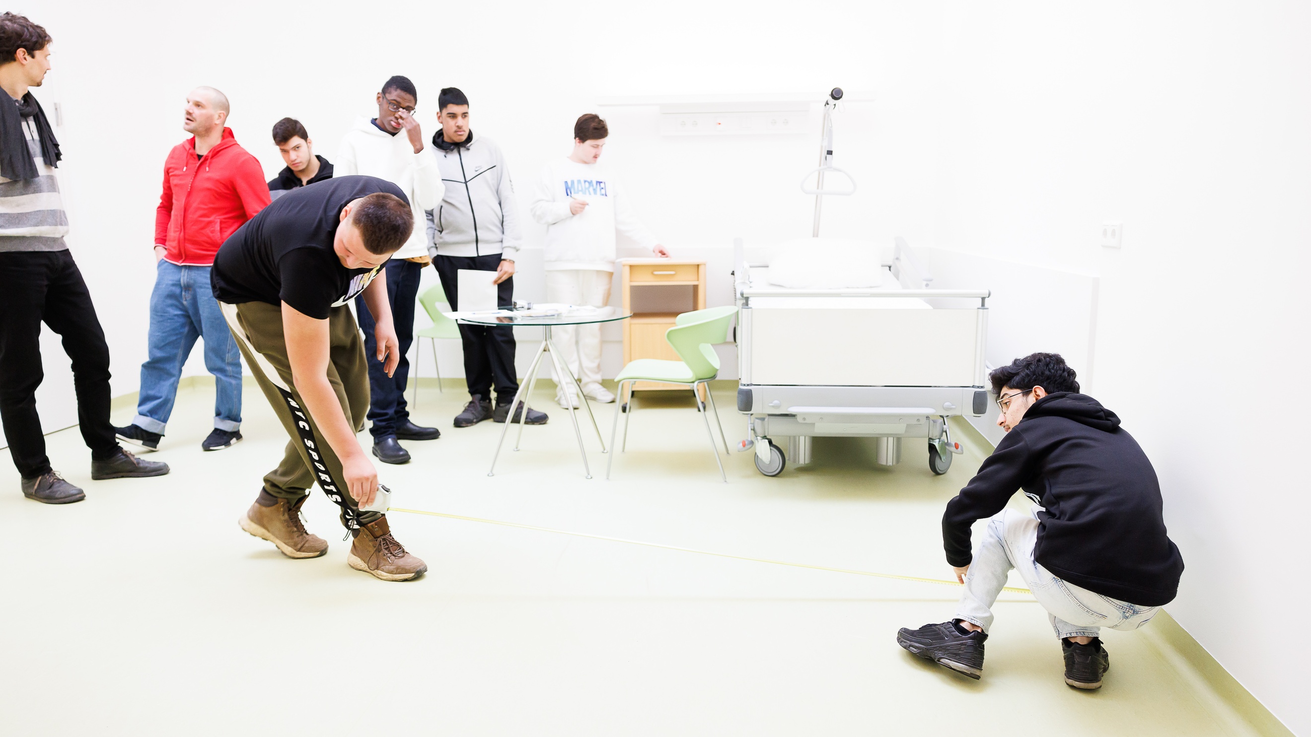In einem Raum mit einem Krankenhausbett stehen mehrere Personen. Zwei Personen messen mit Maßband eine Fläche am Boden