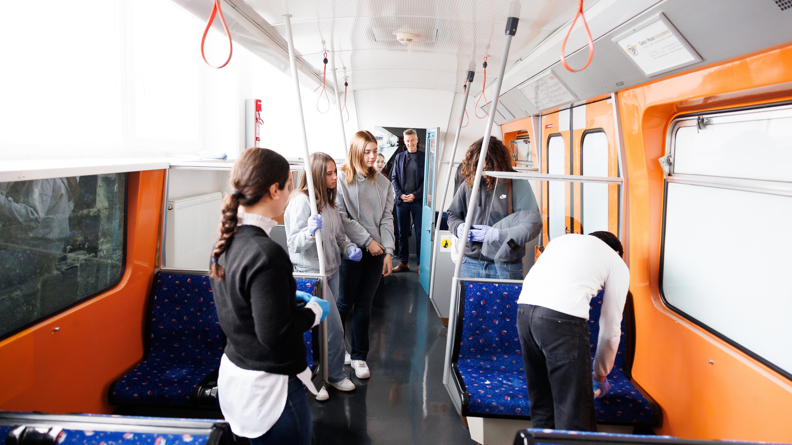 In einer leeren, stehenden U-Bahn stehen mehrere Personen. Ein paar der Personen tragen Handschuhe