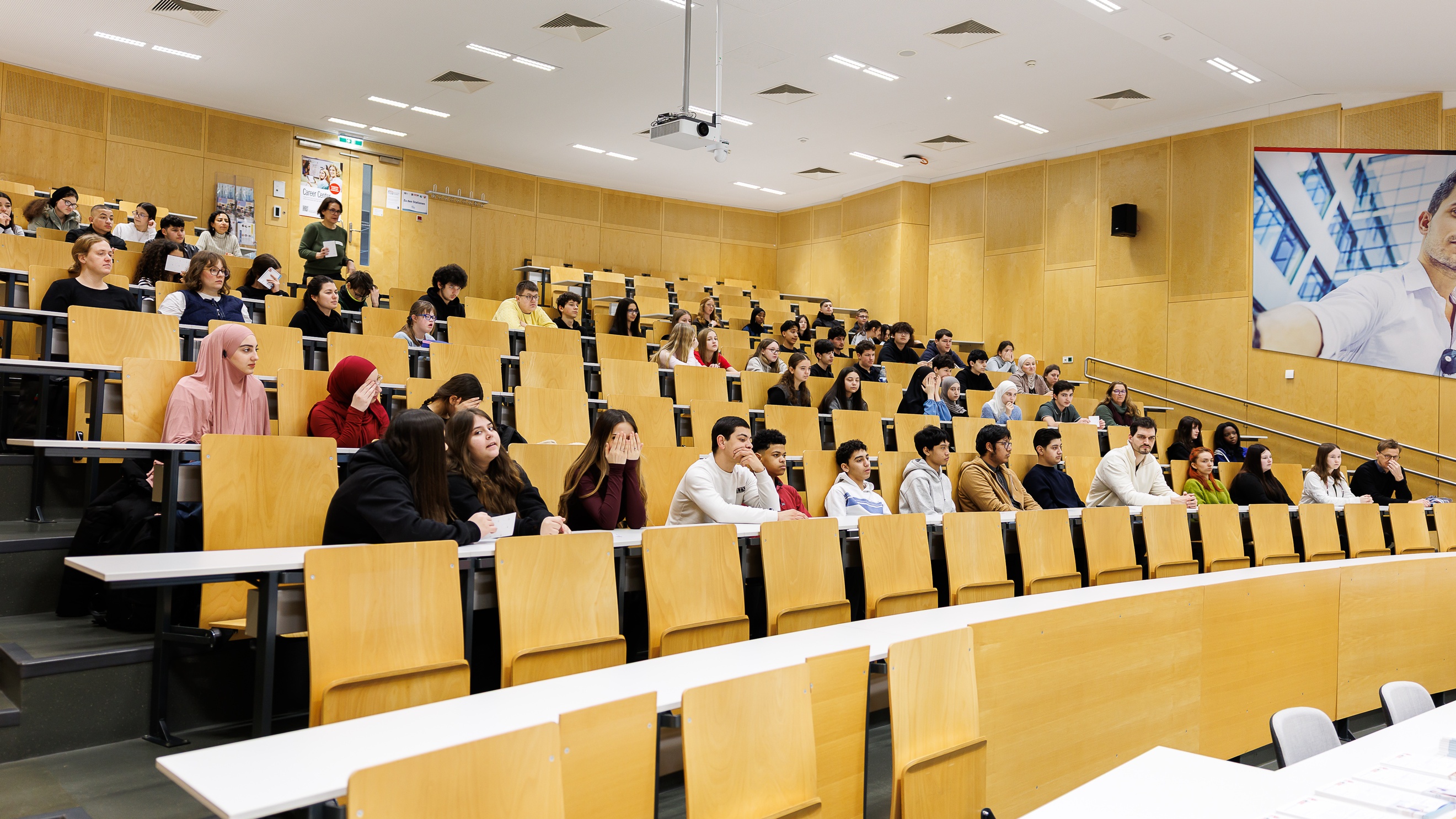 In einem Raum mit aufsteigenden Sitzreihen sitzen hinter den Tischen jugendliche Personen. 